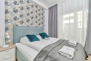 A neatly made bed with a grey blanket and two teal pillows, a bedside table with a lamp, and a wall with a leaf-pattern wallpaper.