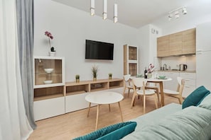 A modern living room with a flat-screen TV, a dining area with a round table and chairs, and a kitchen with wooden cabinets and a kettle.