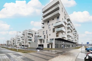 Un immeuble résidentiel moderne à plusieurs étages, doté de balcons, d’un passage piéton et d’un espace de stationnement pour vélos.