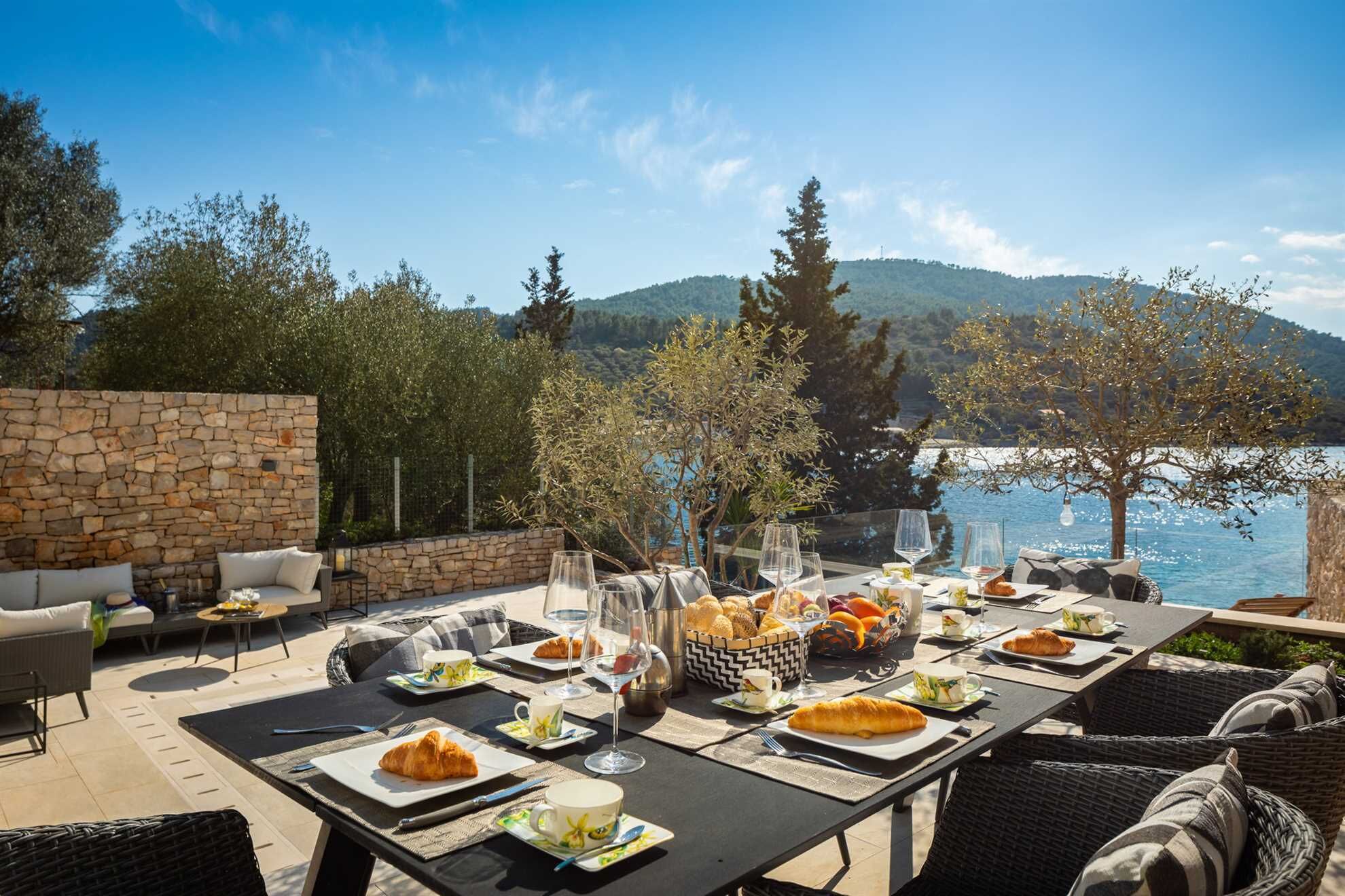 An outdoor dining area with stone walls, wicker furniture, and a scenic view of a lake and mountains.