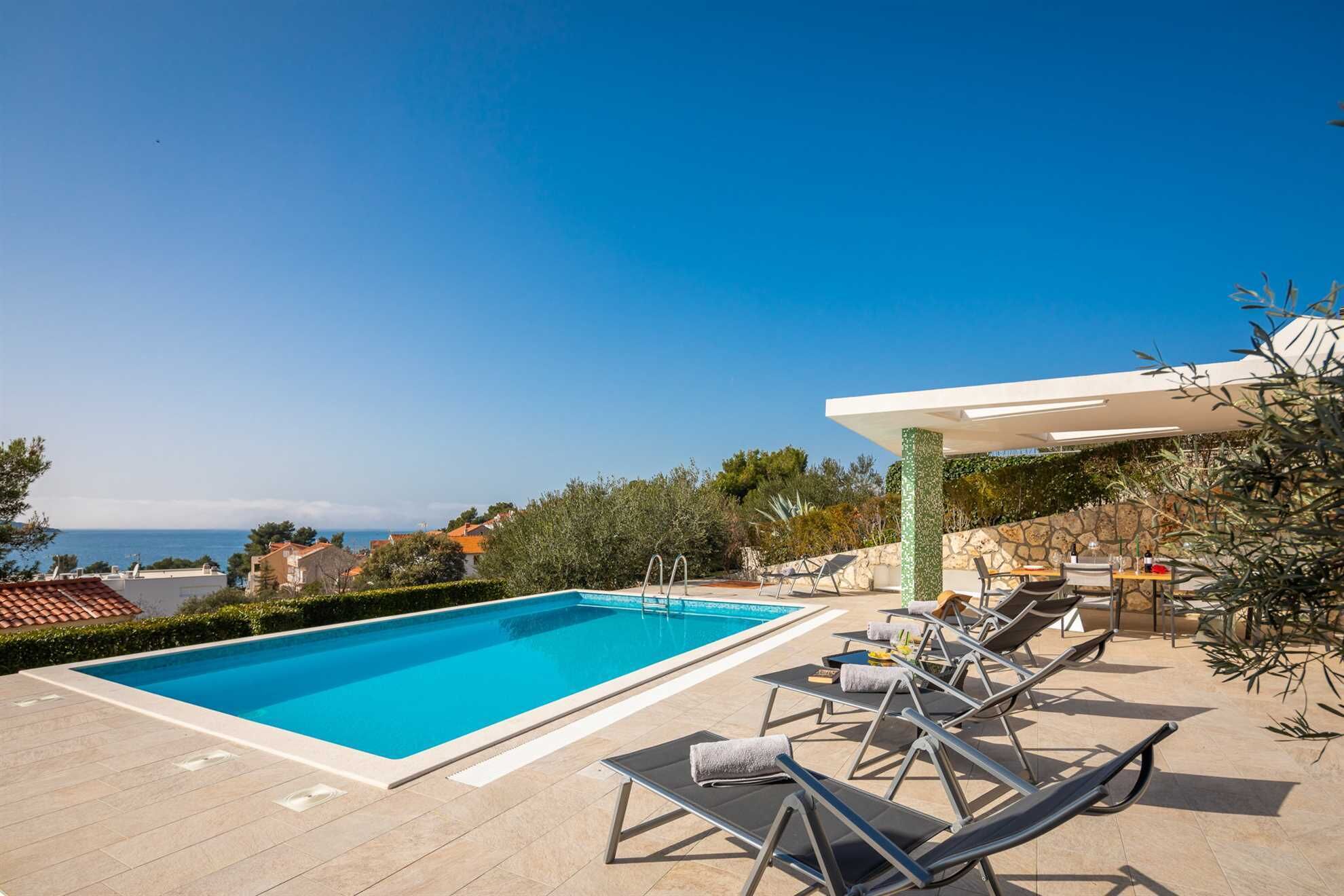 A swimming pool with lounge chairs and a table, surrounded by a stone wall and greenery, with a view of the sea and buildings in the background.