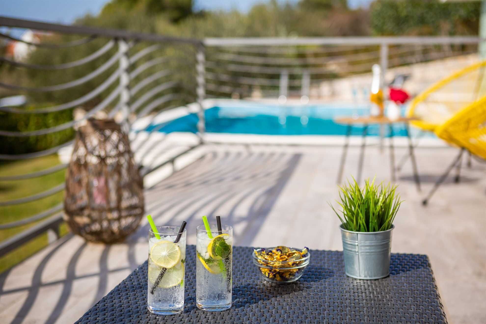 A poolside table with two glasses of iced drinks, a small potted plant, and a bowl of dried flowers.