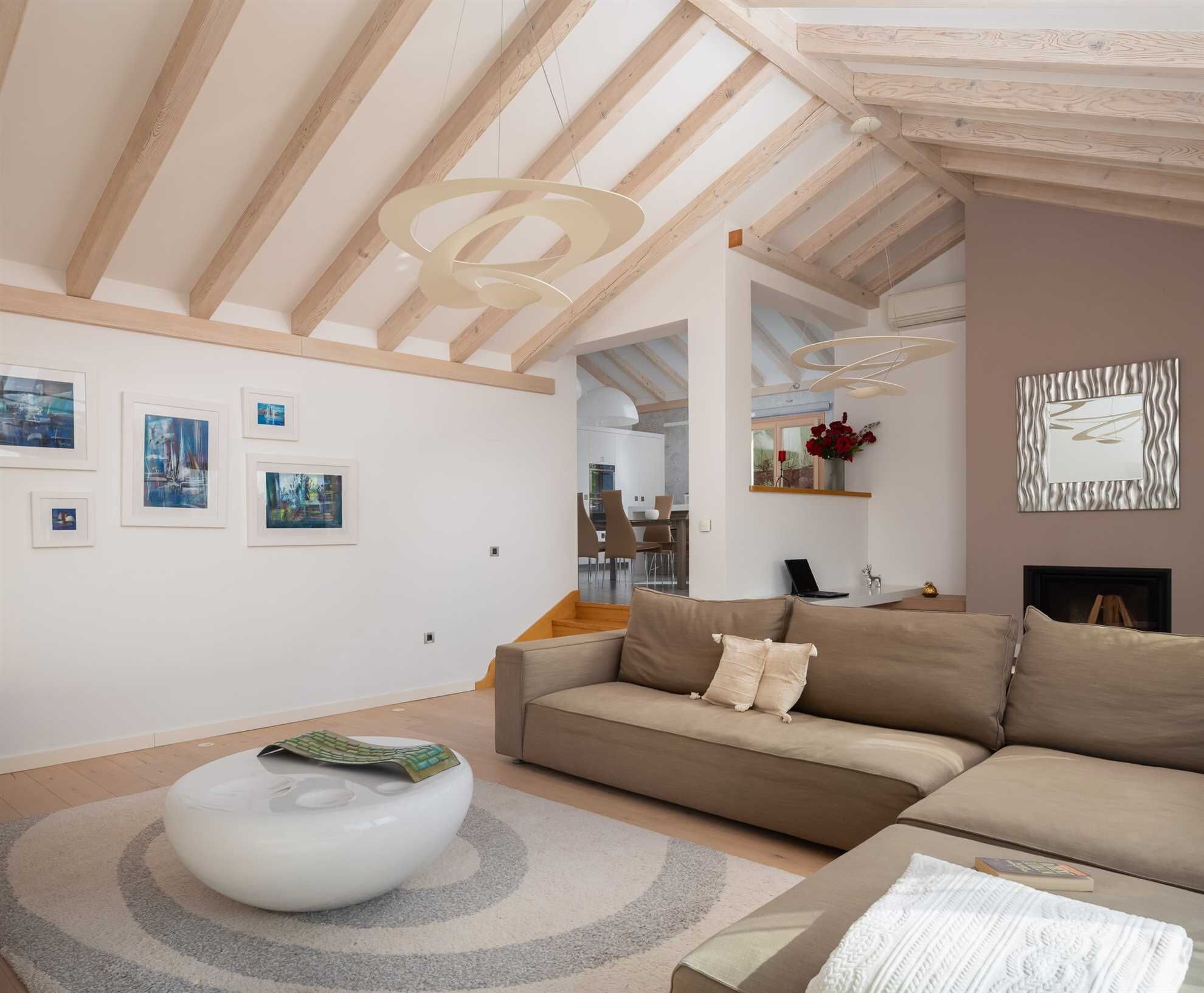 A modern living room with a high ceiling, wooden beams, a beige sofa, a white ottoman, and framed artwork on the walls.