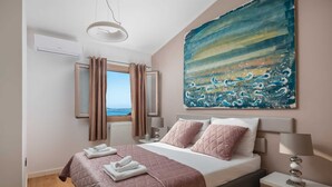 Een slaapkamer met een bed, nachtkastje, lamp, schilderij en raam met gordijnen.