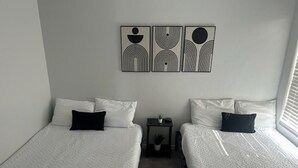 Een kamer met twee bedden, wit beddengoed, een kleine tafel met een plant en abstracte wanddecoratie.