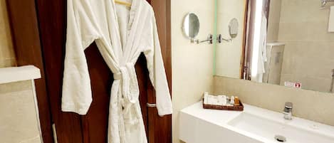 Un baño con puerta de madera, una bata blanca colgada en la puerta, un espejo grande y un lavabo blanco con artículos de aseo.
