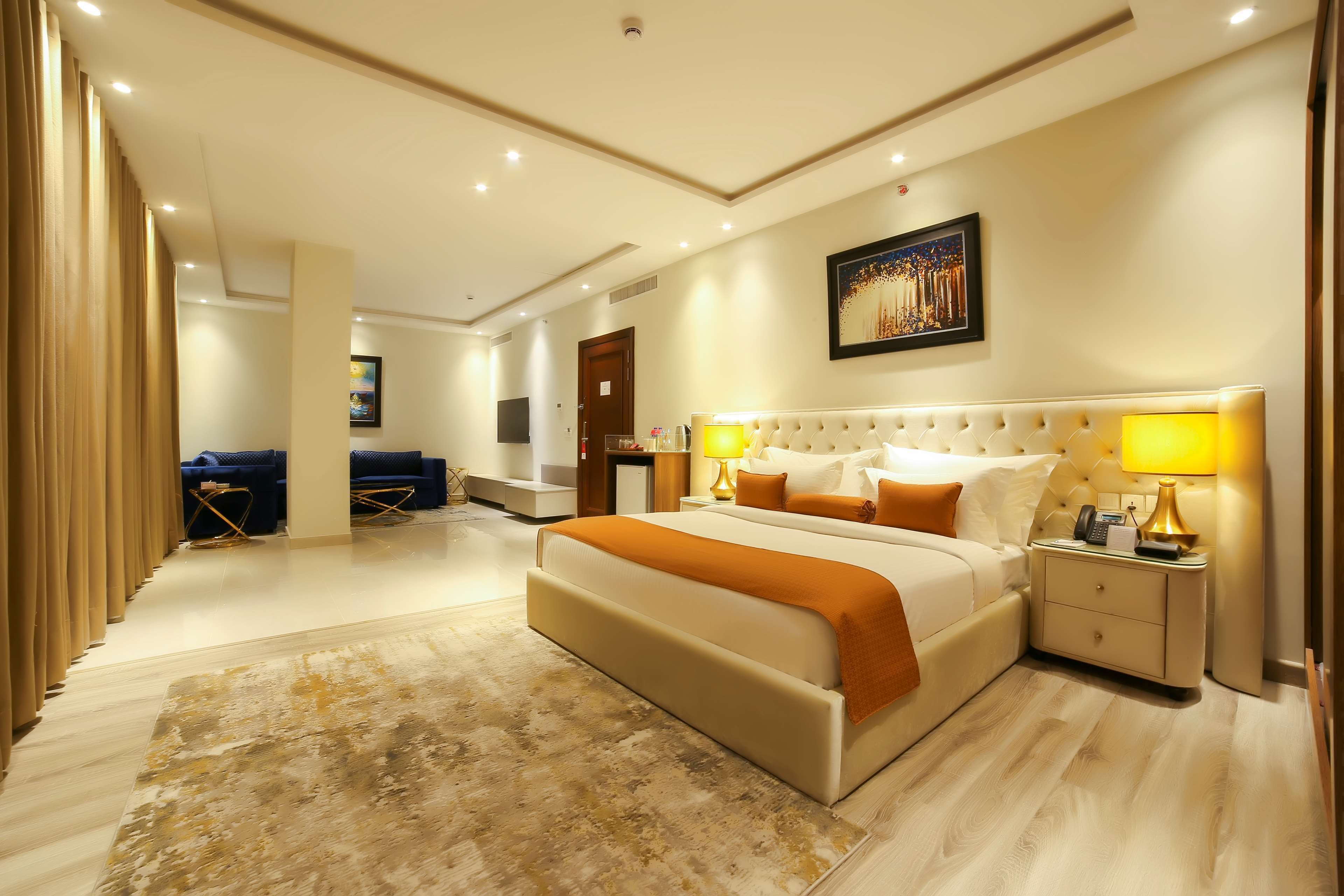 A spacious hotel room with a large bed, a bedside table with lamps, a wall-mounted TV, and a large window.