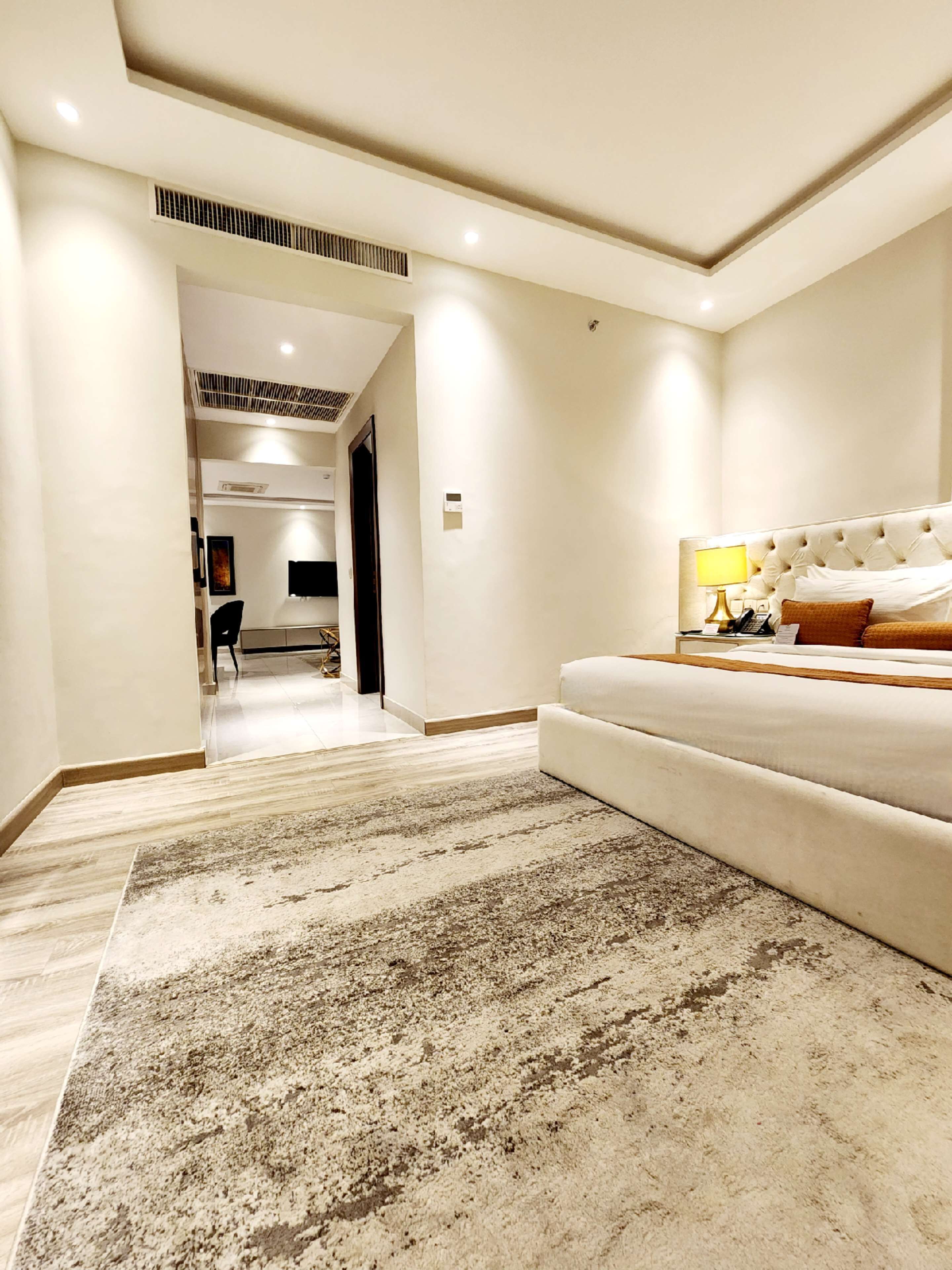A spacious bedroom with a large bed, a bedside table, and a lamp. There is a carpeted floor and a hallway leading to another room.