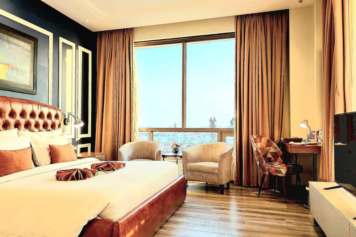 Habitación de hotel con una cama grande, dos sillones, un escritorio y un ventanal con cortinas.