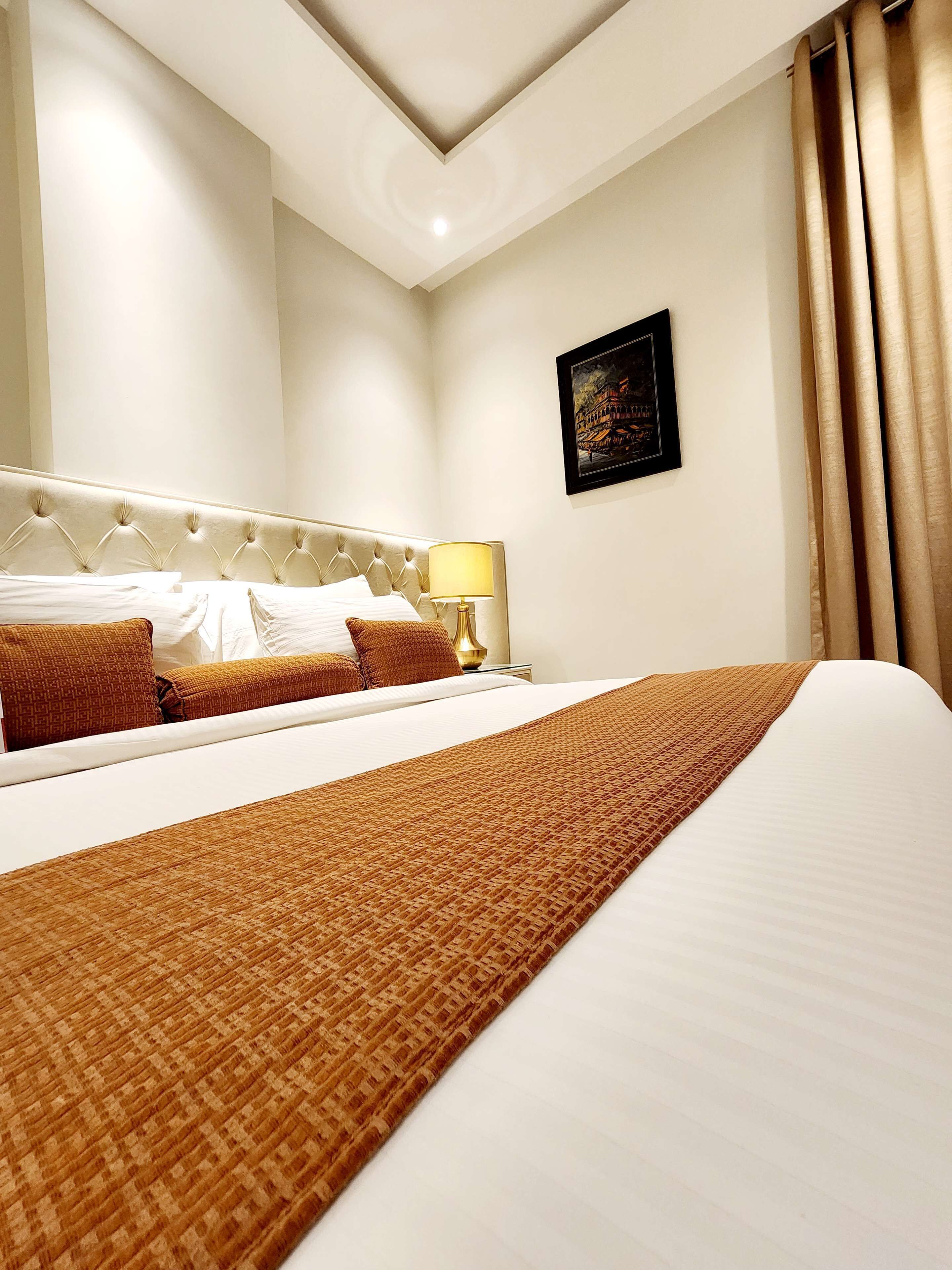 A hotel room with a large bed, a headboard, a bedside table with a lamp, and a framed picture on the wall.