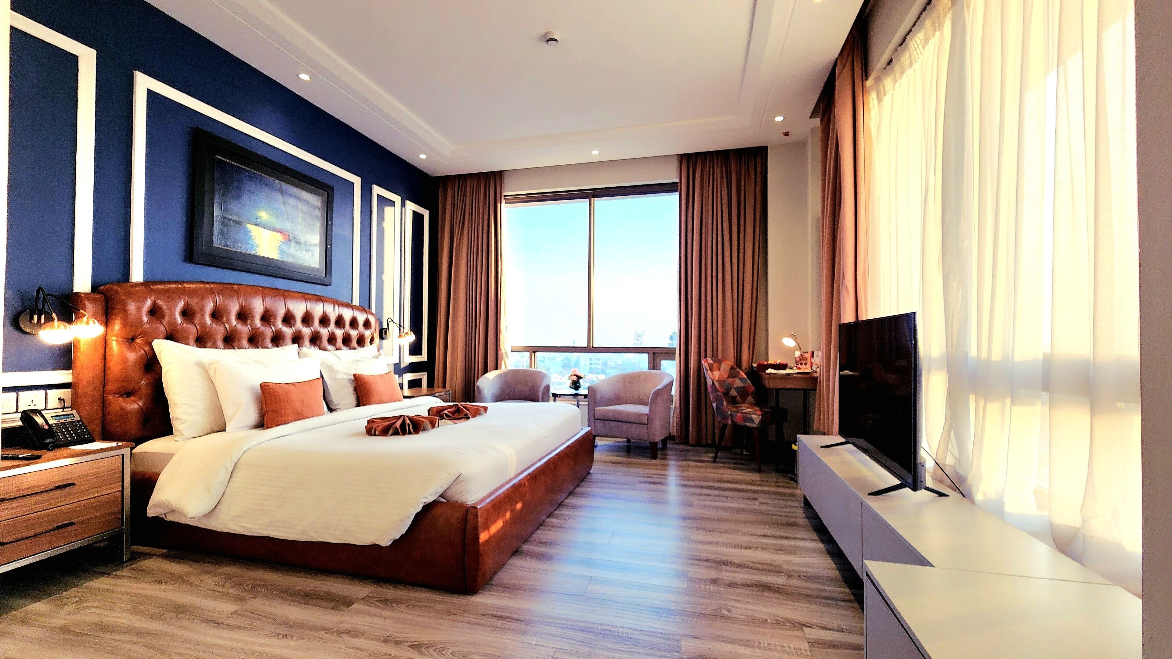A spacious hotel room with a large bed, a desk with a TV, and a view of the city through large windows.