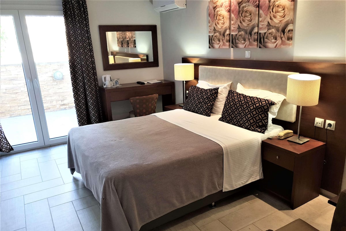 A hotel room with a bed, a bedside table with a lamp, a chair, a desk with a mirror, and a large window.