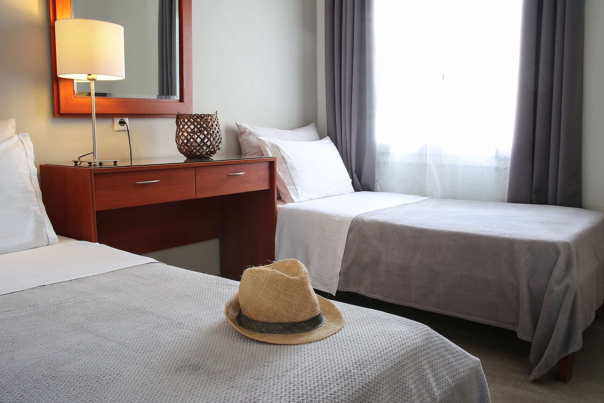 A hotel room with two beds, a wooden nightstand, a lamp, a mirror, and a window with curtains.