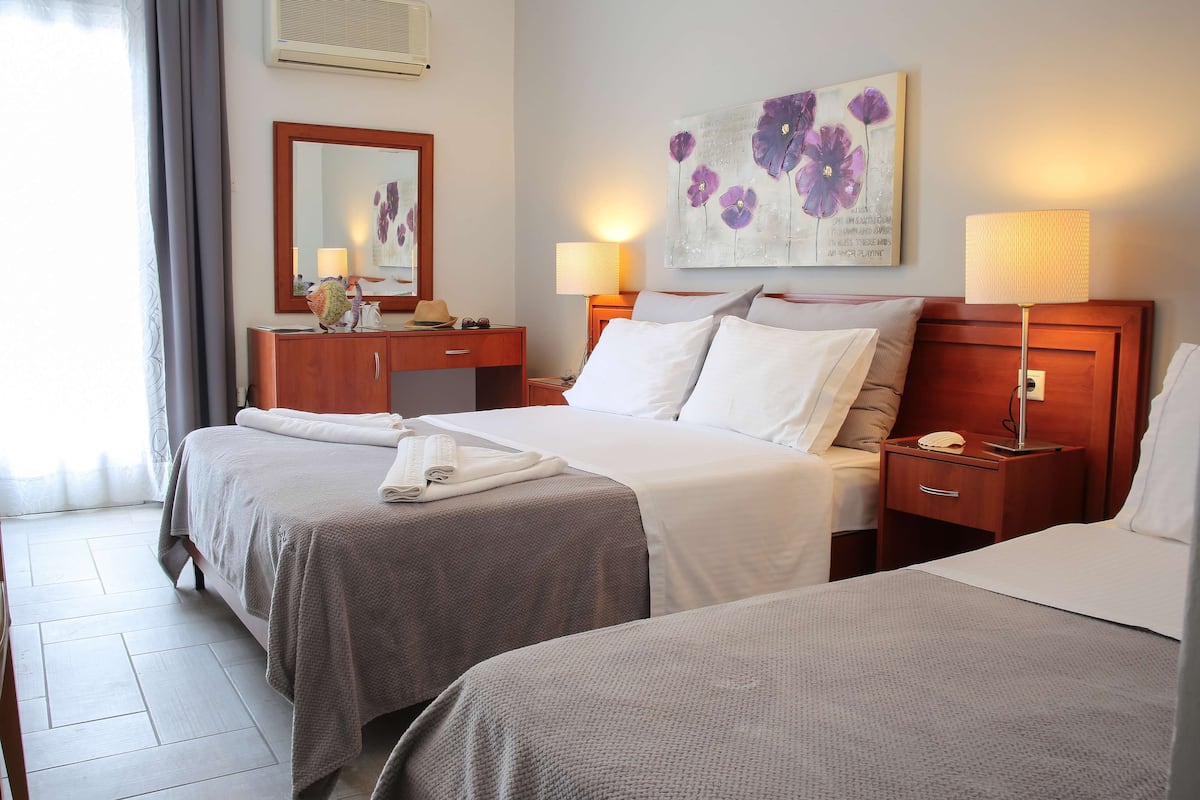 A hotel room with two beds, a wooden headboard, a bedside table with a lamp, a mirror, and a painting of flowers on the wall.