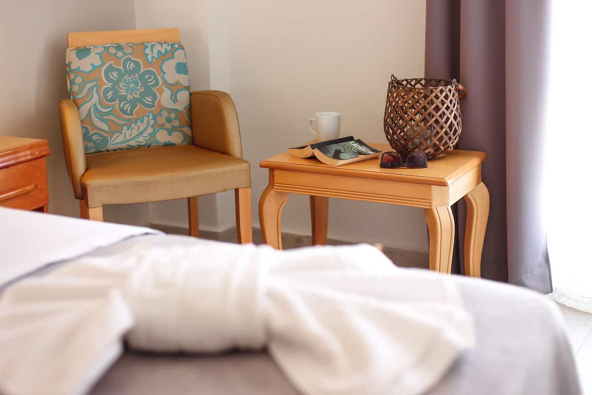 A hotel room with a bed, a chair with a patterned cushion, a small wooden table with a white mug, a book, and a woven basket.