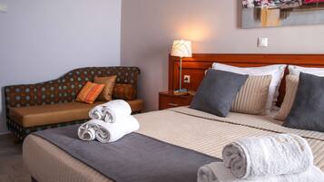 A neatly made bed with a grey blanket and two rolled white towels, a brown sofa with patterned cushions, and a bedside table with a lamp.