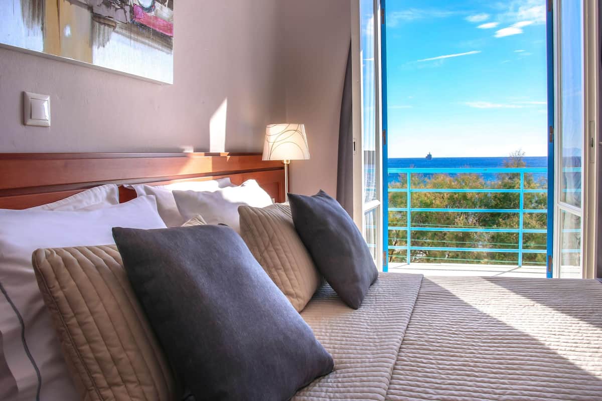 A hotel room with a bed, pillows, a lamp, and a view of the sea through a balcony.