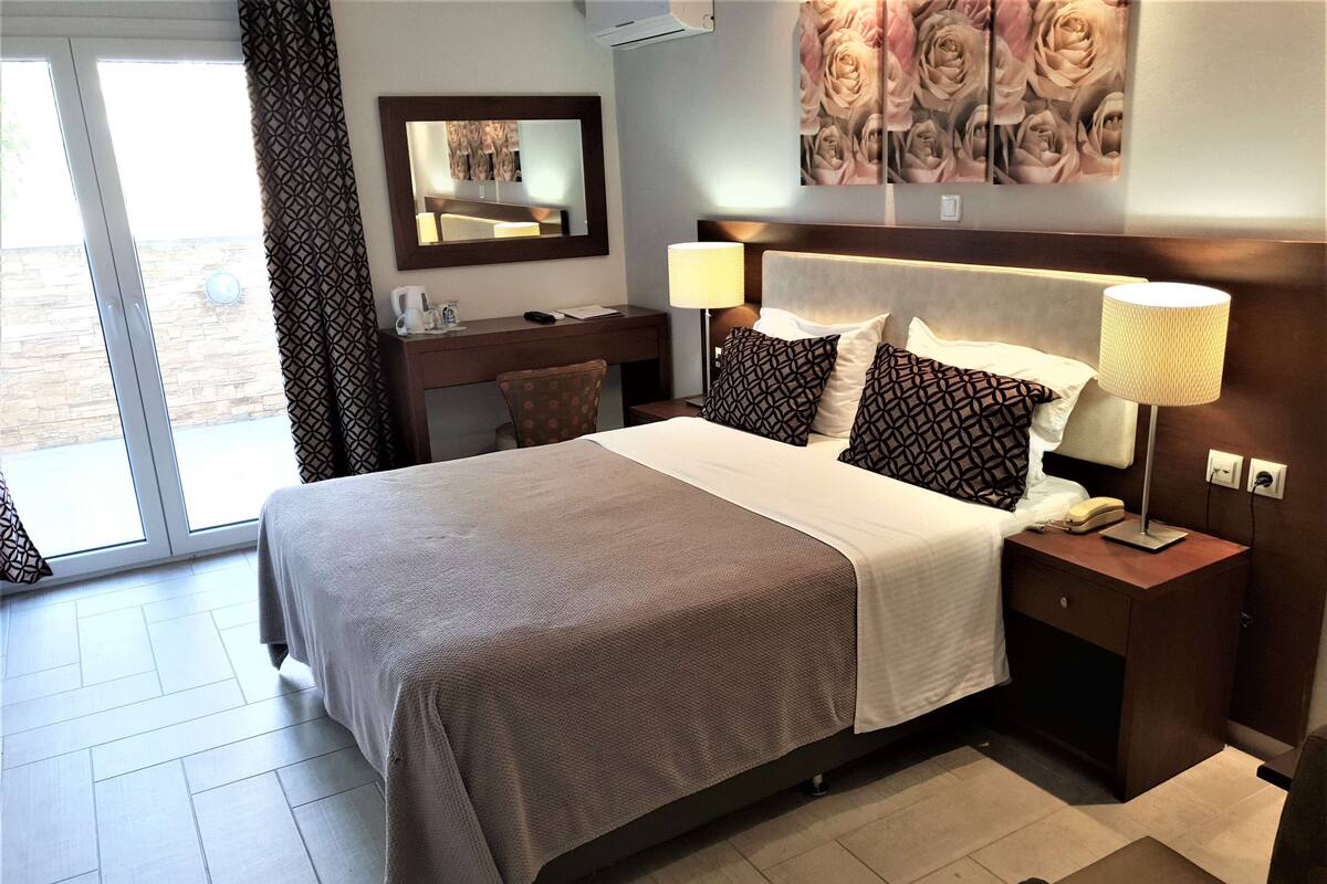 A hotel room with a large bed, a desk with a chair, a bedside table with a lamp, a mirror, and a wall with a floral print.