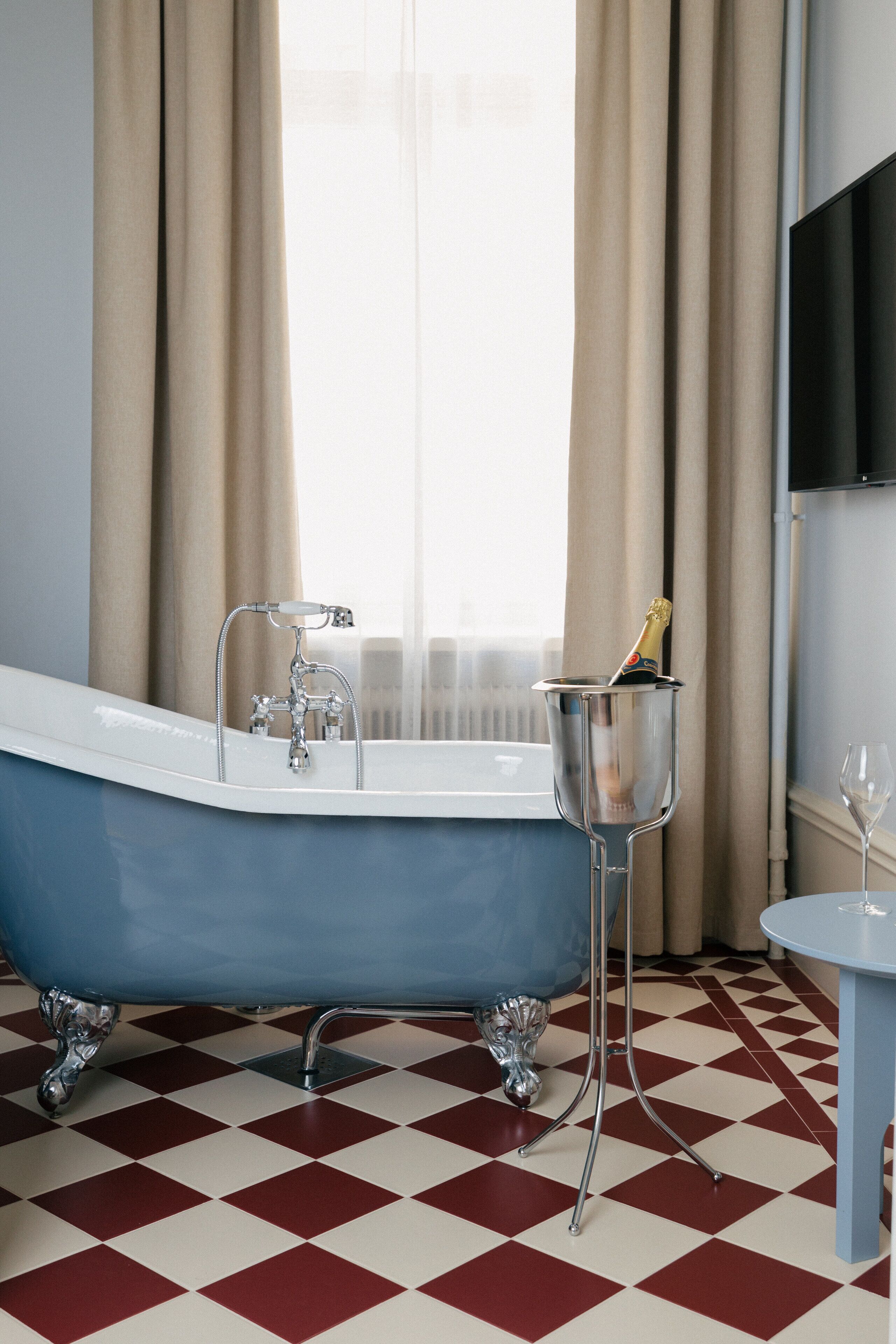 A bathroom with a blue clawfoot bathtub, a champagne bottle in a glass, and a small table with a wine glass.