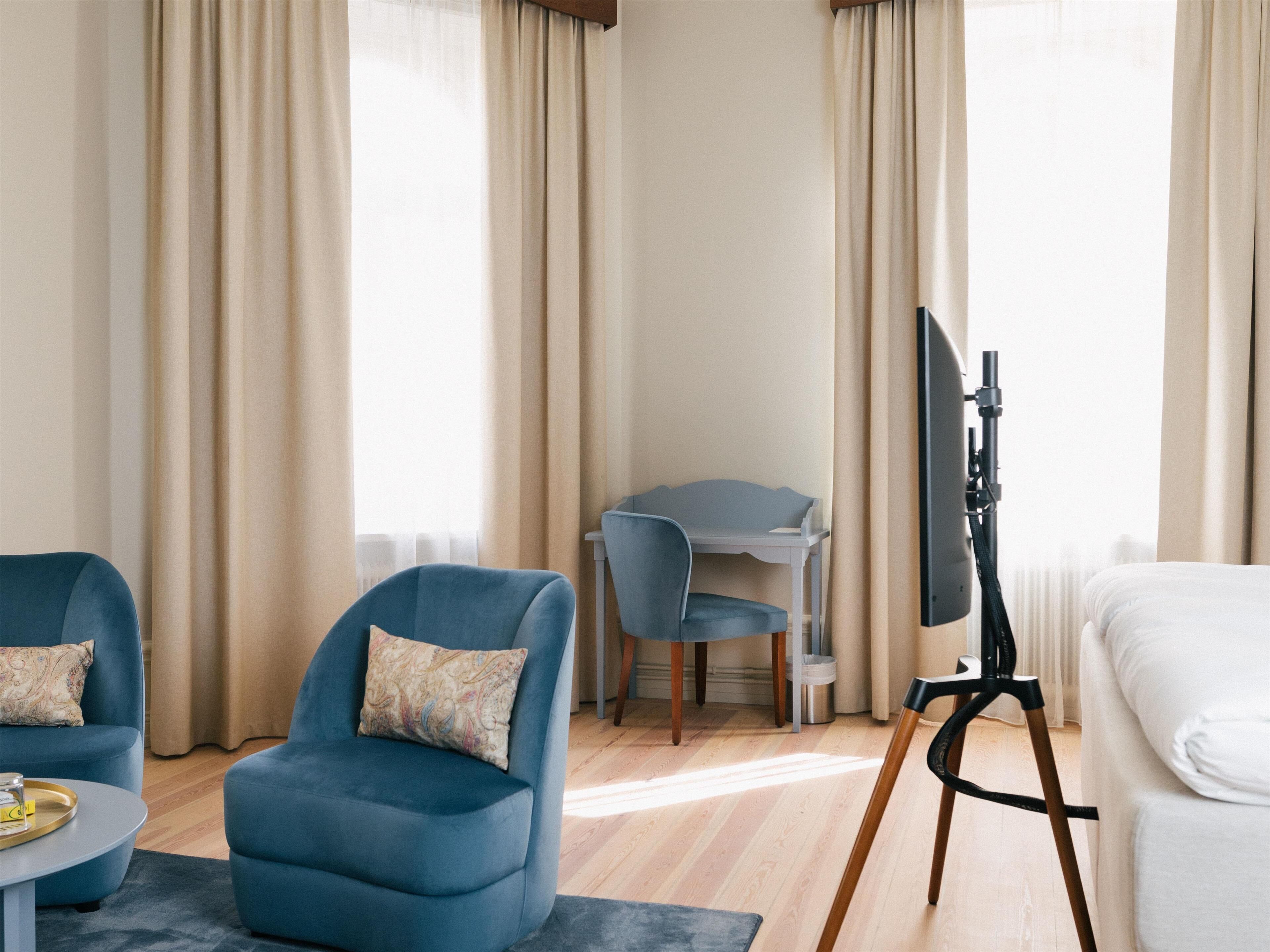 Una habitación de hotel con un sillón azul, un sofá blanco, un escritorio con una computadora y un televisor sobre un trípode.