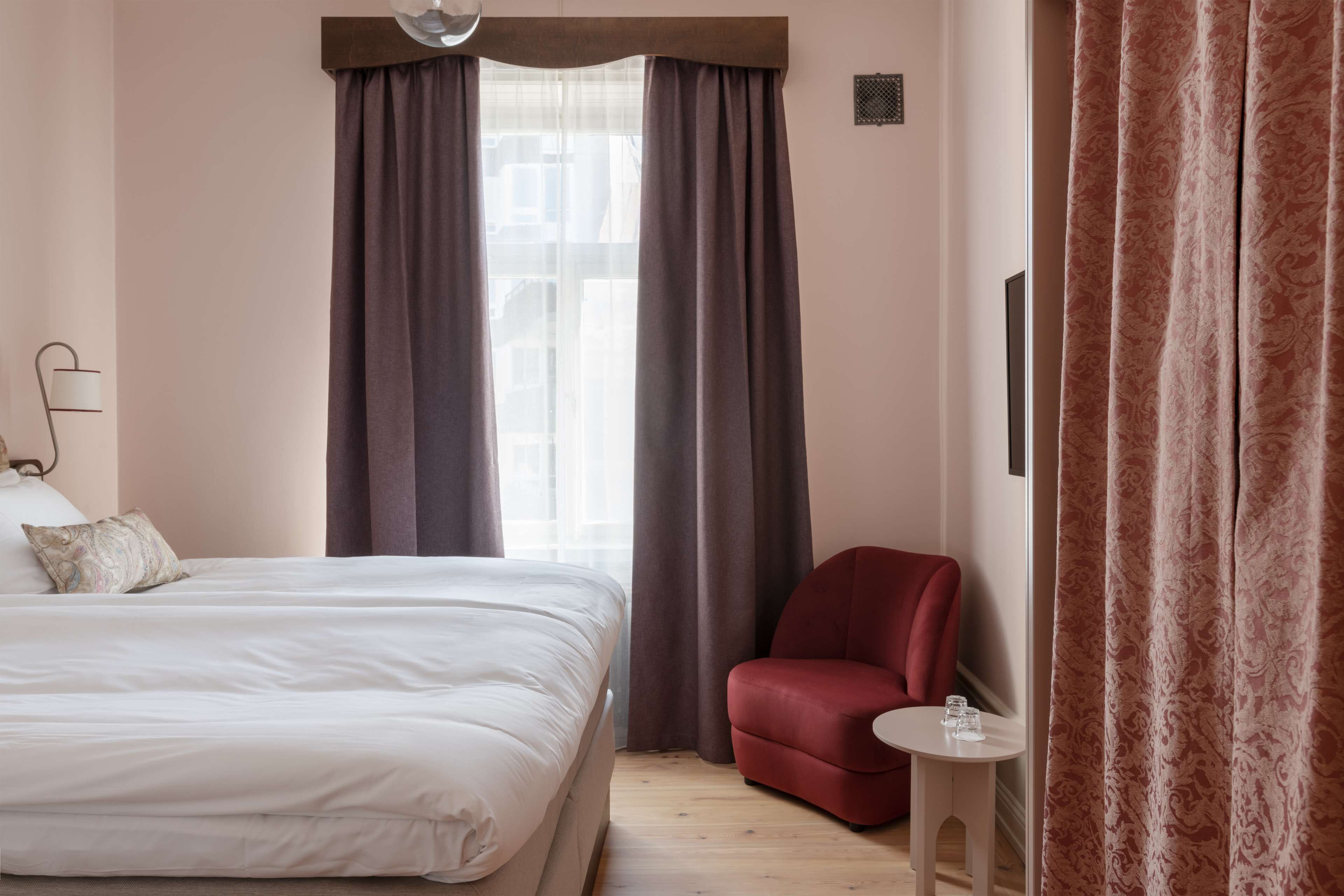 A hotel room with a bed, a red armchair, a small table with a glass, and a window with curtains.