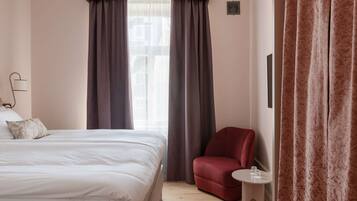 A hotel room with a bed, a red armchair, a small table with a glass, and a window with curtains.