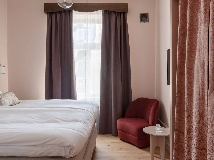 A hotel room with a bed, a red armchair, a small table with a glass, and a window with curtains.