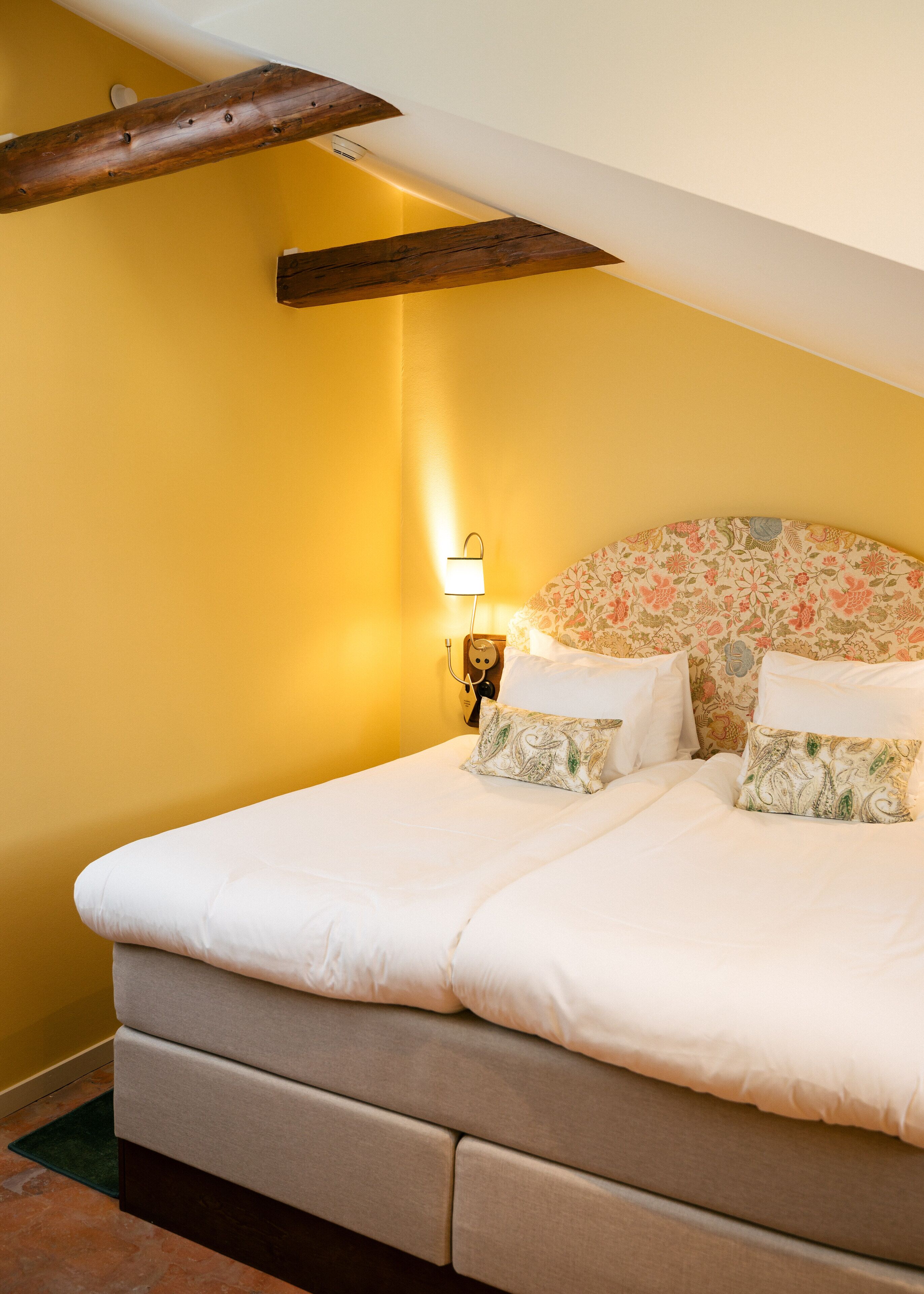 Un dormitorio con cama, cabecera de flores, lámpara de noche y vigas de madera en el techo.