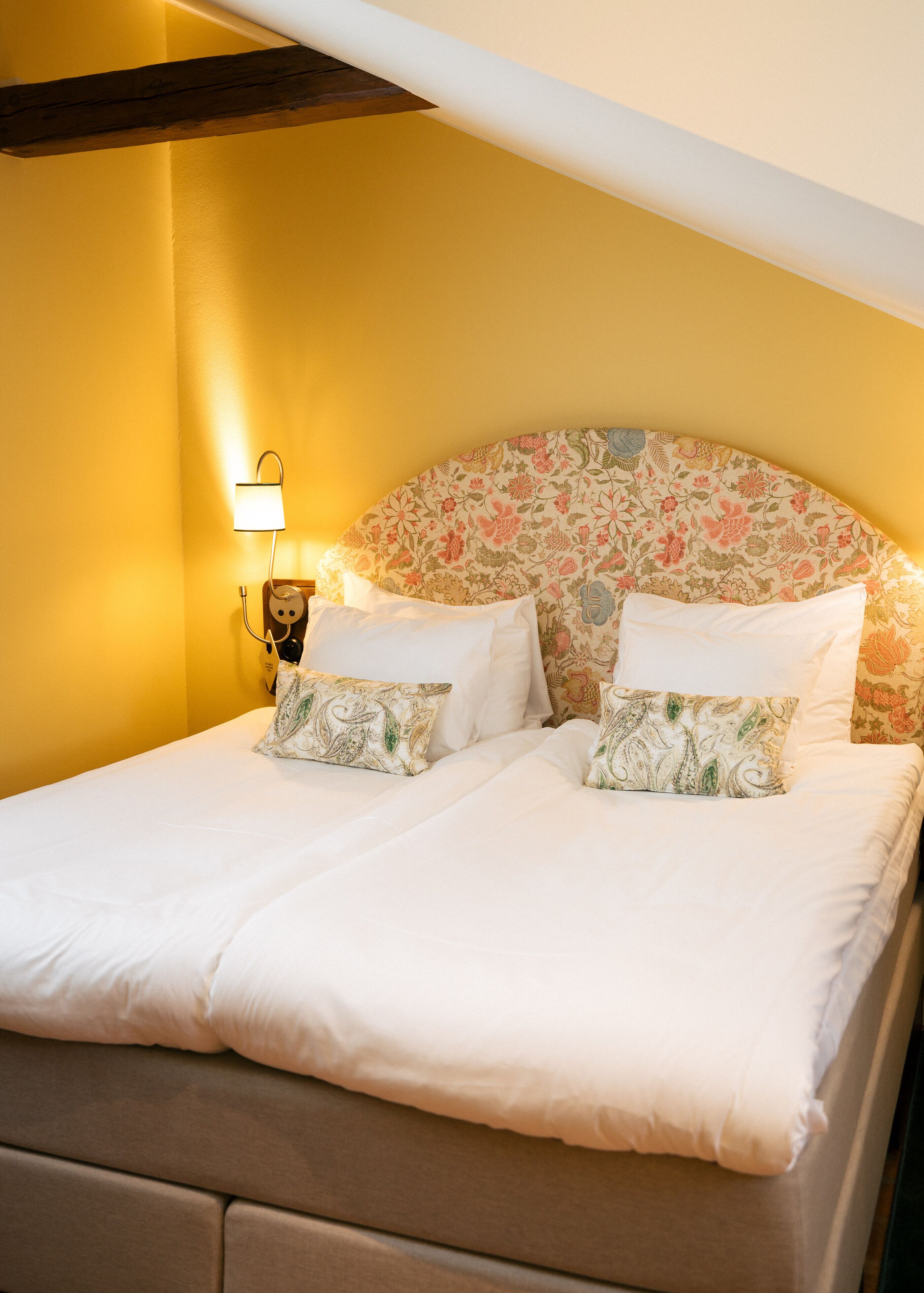 A bed with white linens and floral-patterned pillows, a wall-mounted lamp, and a wooden beam structure.