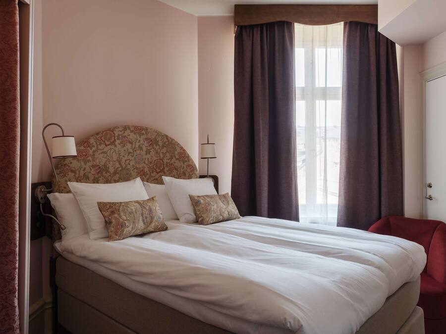 Ein ordentlich bezogenes Bett mit weißen Leinen und gemusterten Kissen, ein roter Sessel und ein großes Fenster mit Vorhängen.