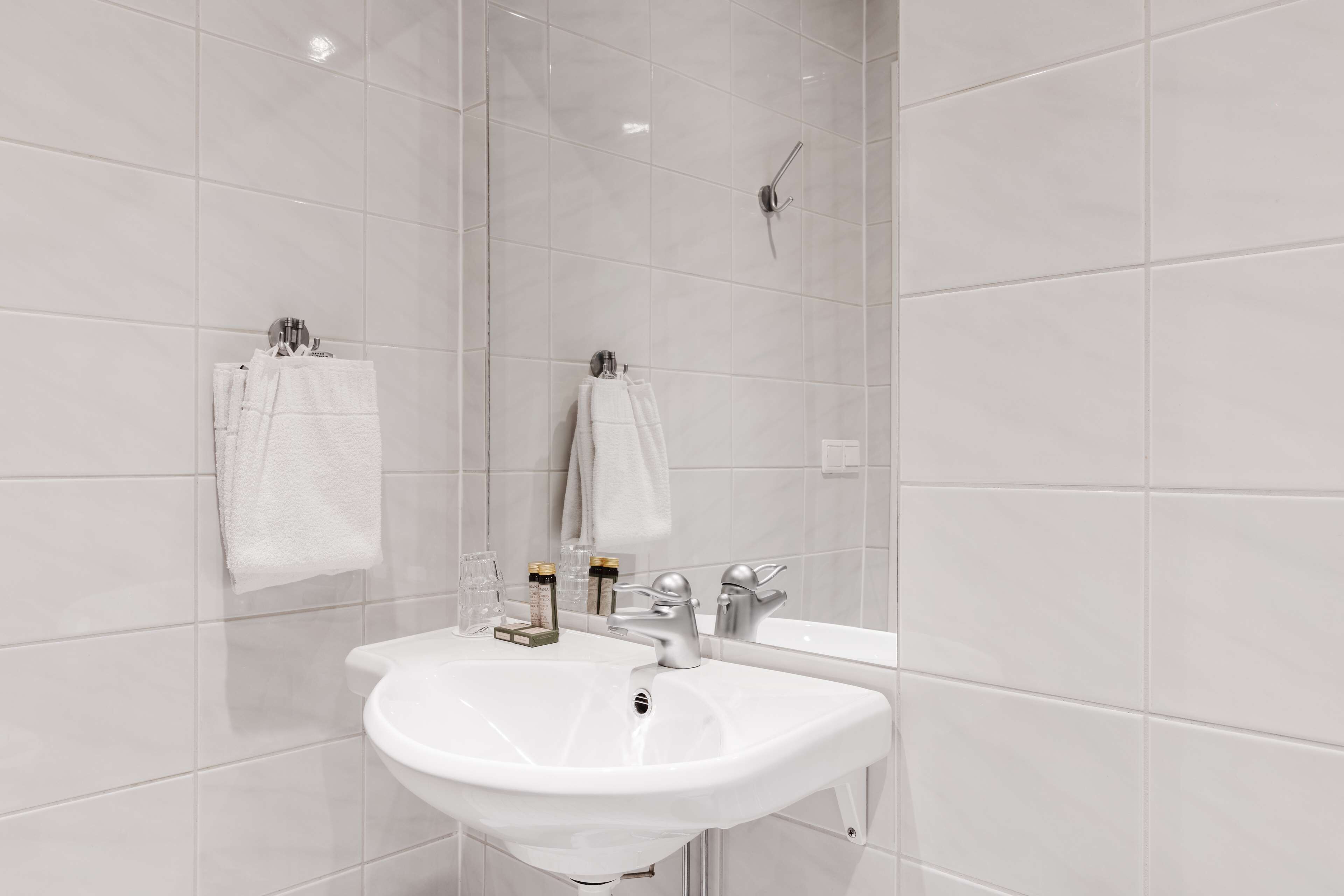 A modern bathroom with white tiles, a sink, a mirror, and two towels hanging on hooks.