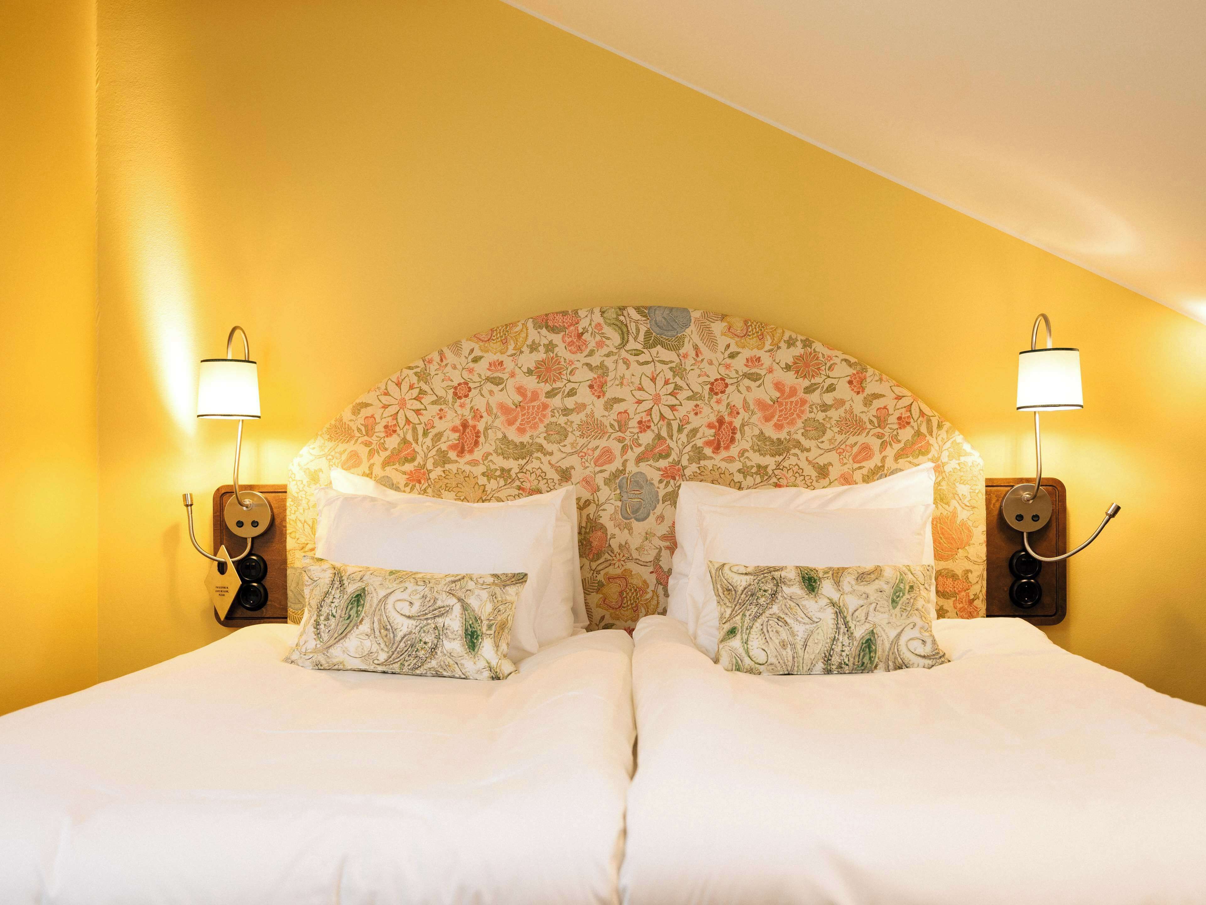 Cama doble con cabecera floral y dos lámparas de noche, frente a una pared amarilla.