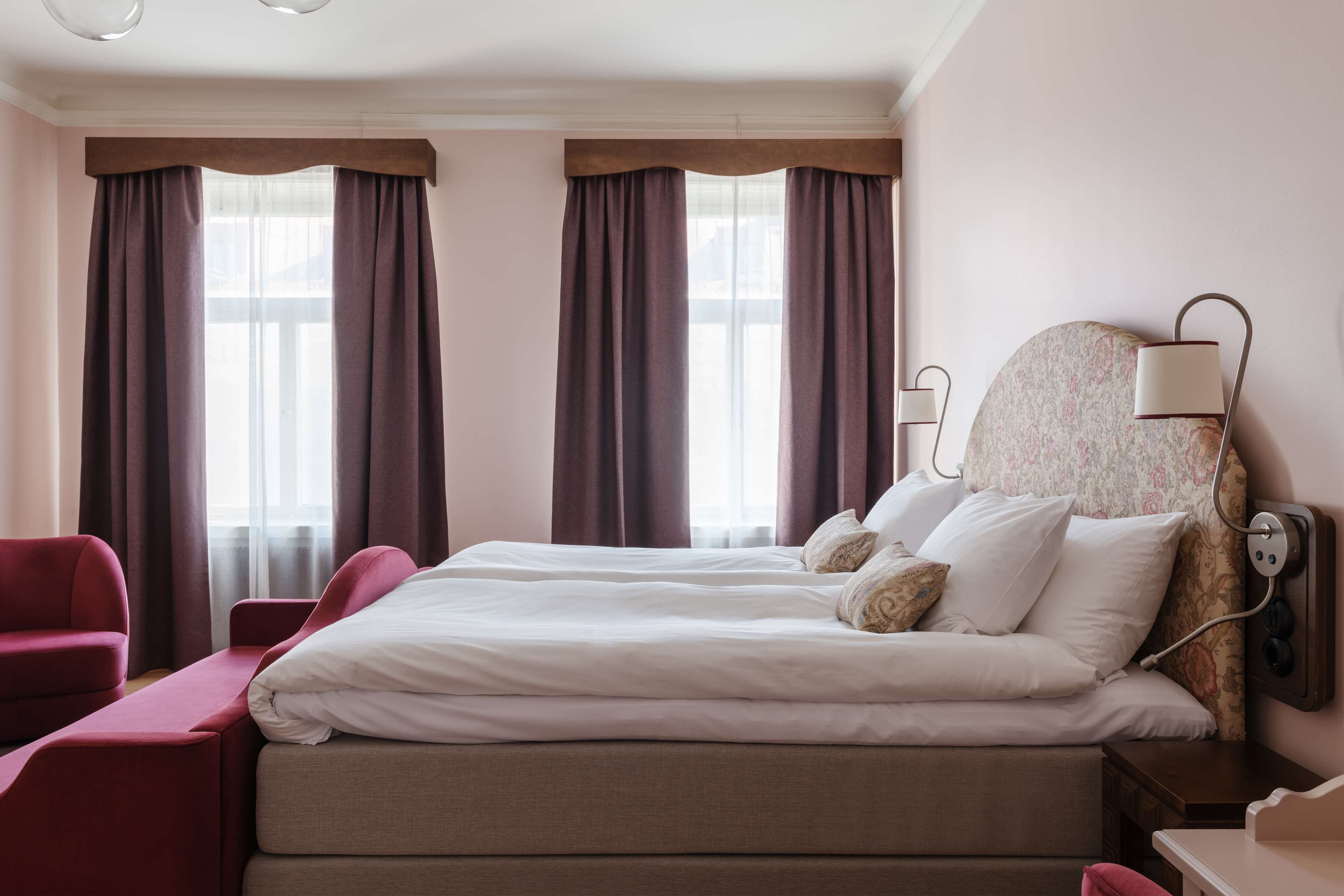 Una camera d'albergo con un grande letto, una poltrona rossa, un comodino con una lampada e una finestra con tende.
