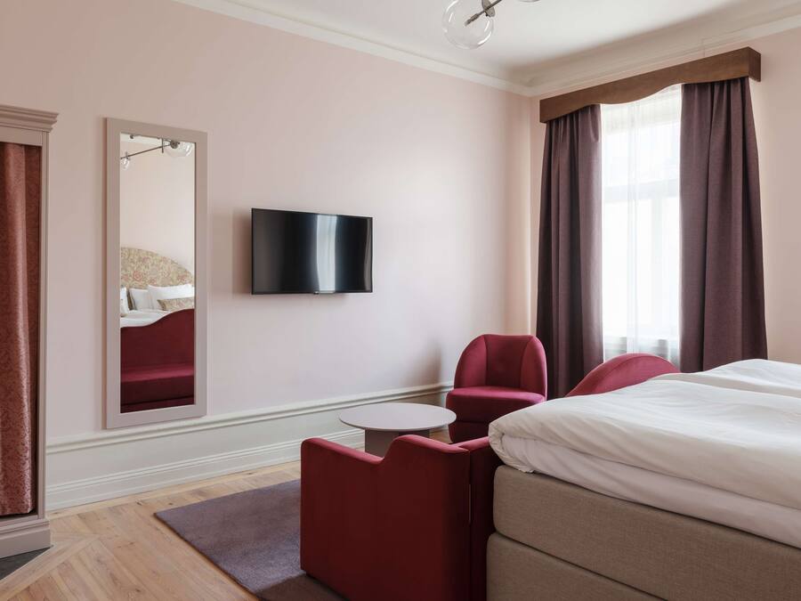A hotel room with a bed, a TV, a red armchair, and a small table.