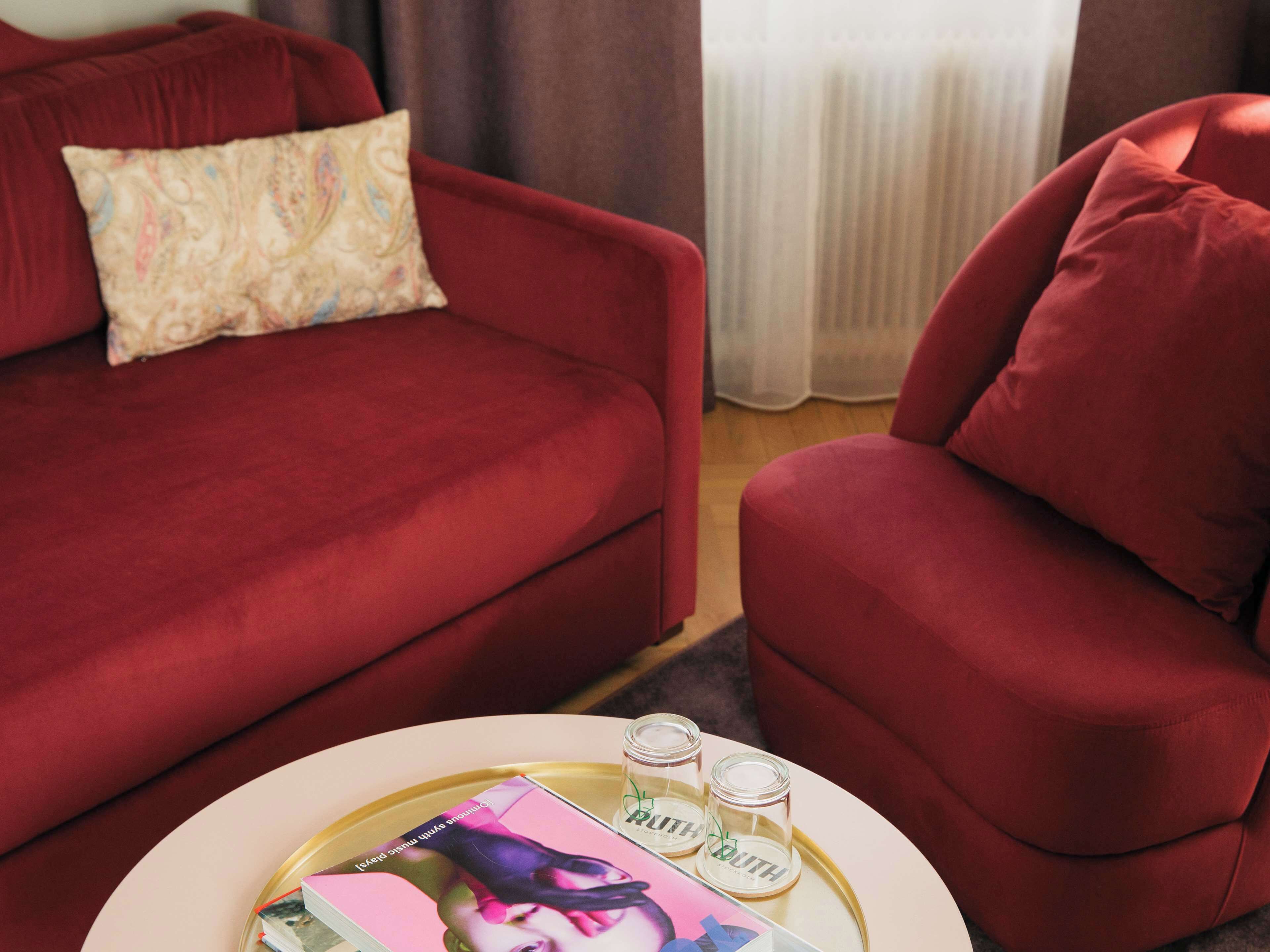 A red sofa and armchair with a decorative pillow, a round table with a magazine and two glass jars, and a window with sheer curtains.