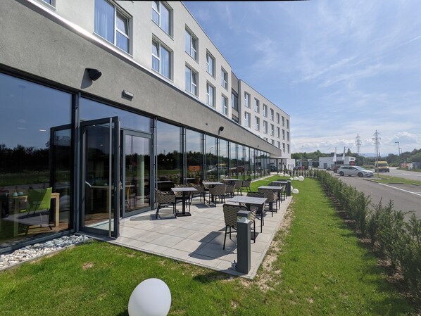 A modern building with a glass facade, outdoor seating area with tables and chairs, and a grassy lawn. - Aiden by Best Western Stadtgut Hotel Steyr (Steyr)