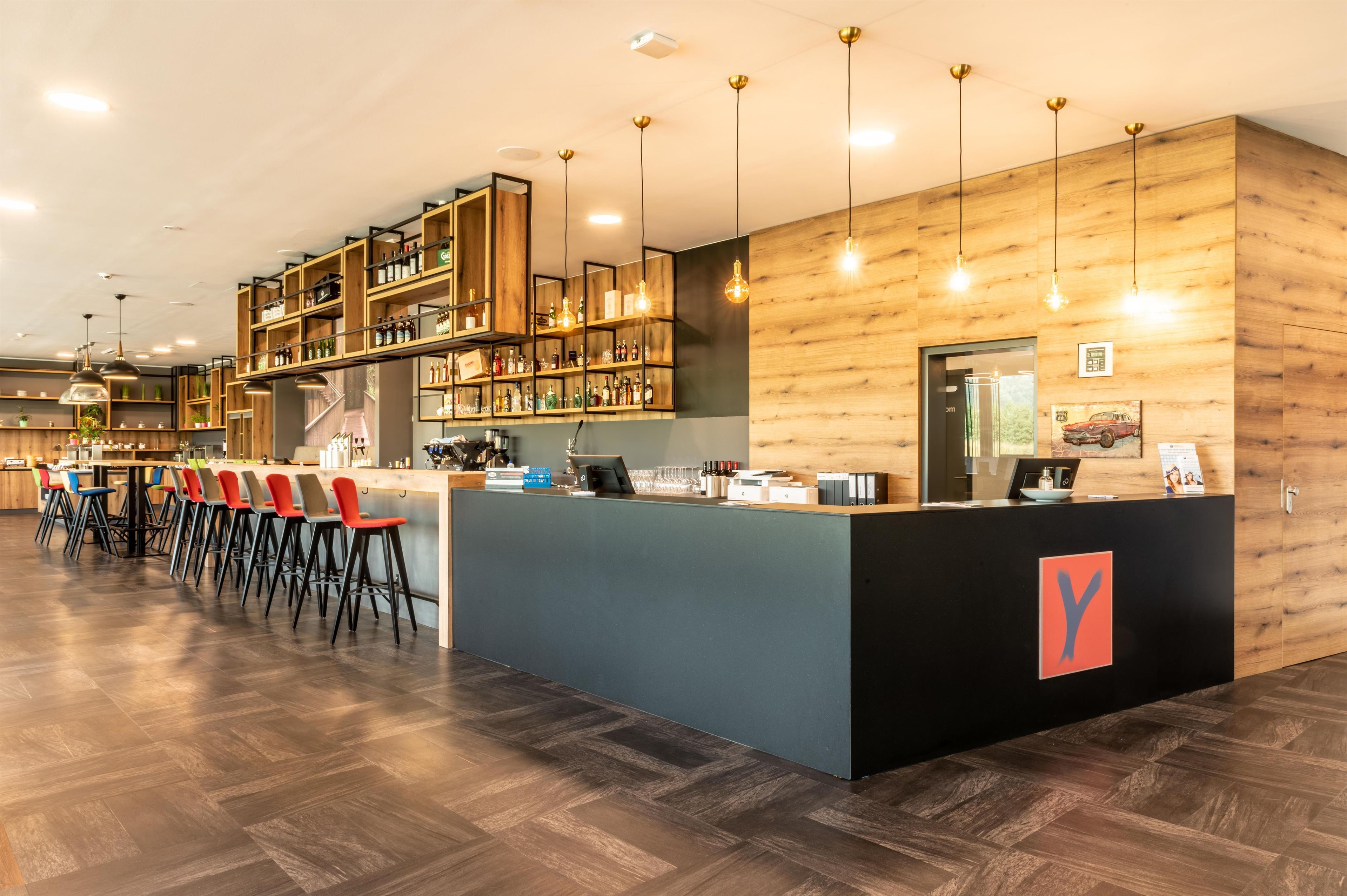 A modern bar area with a wooden bar counter, multiple shelves with bottles, and a seating area with chairs.