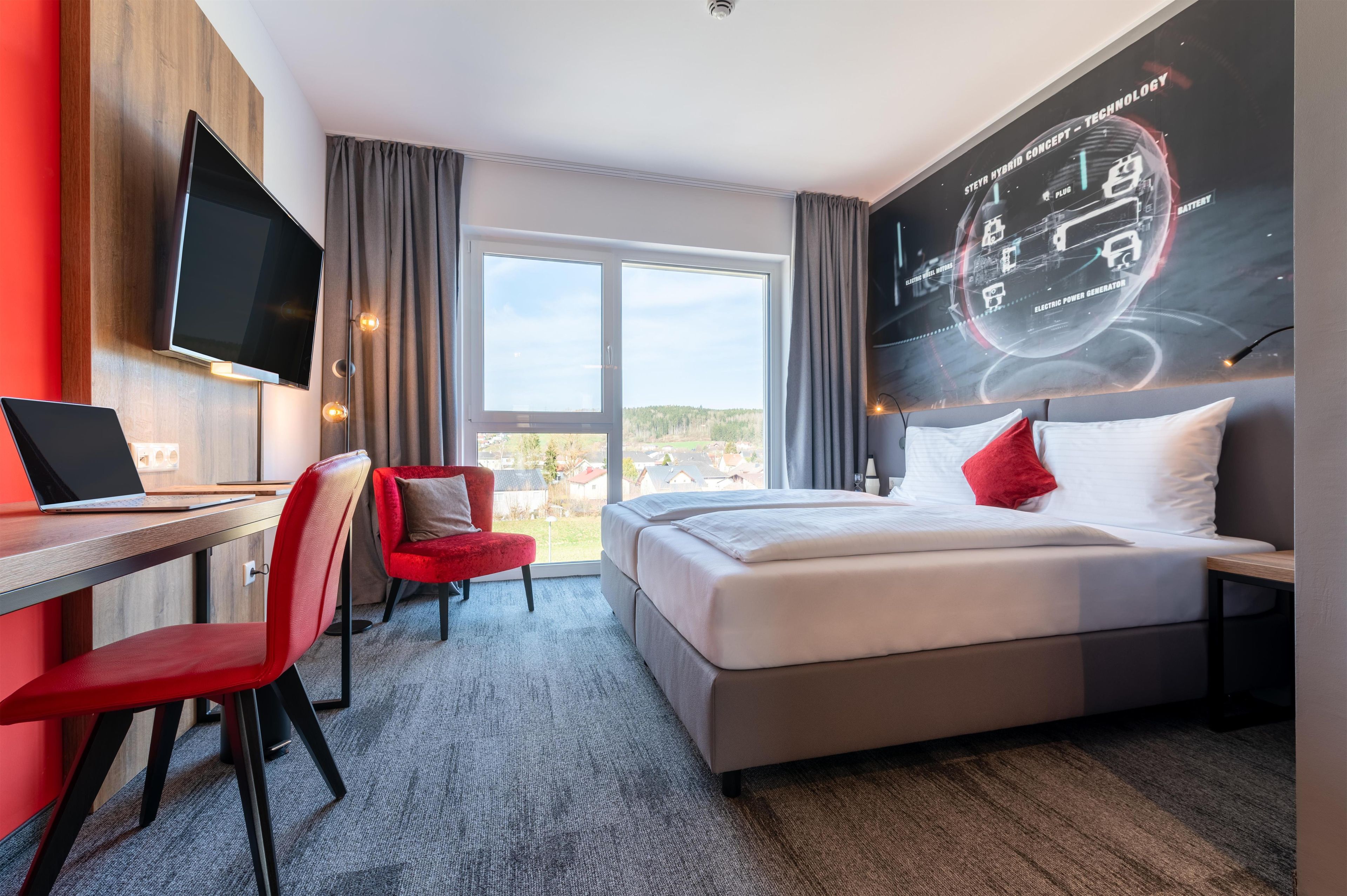 Habitación de hotel con cama, escritorio con una computadora portátil, televisor y un amplio ventanal con cortinas.