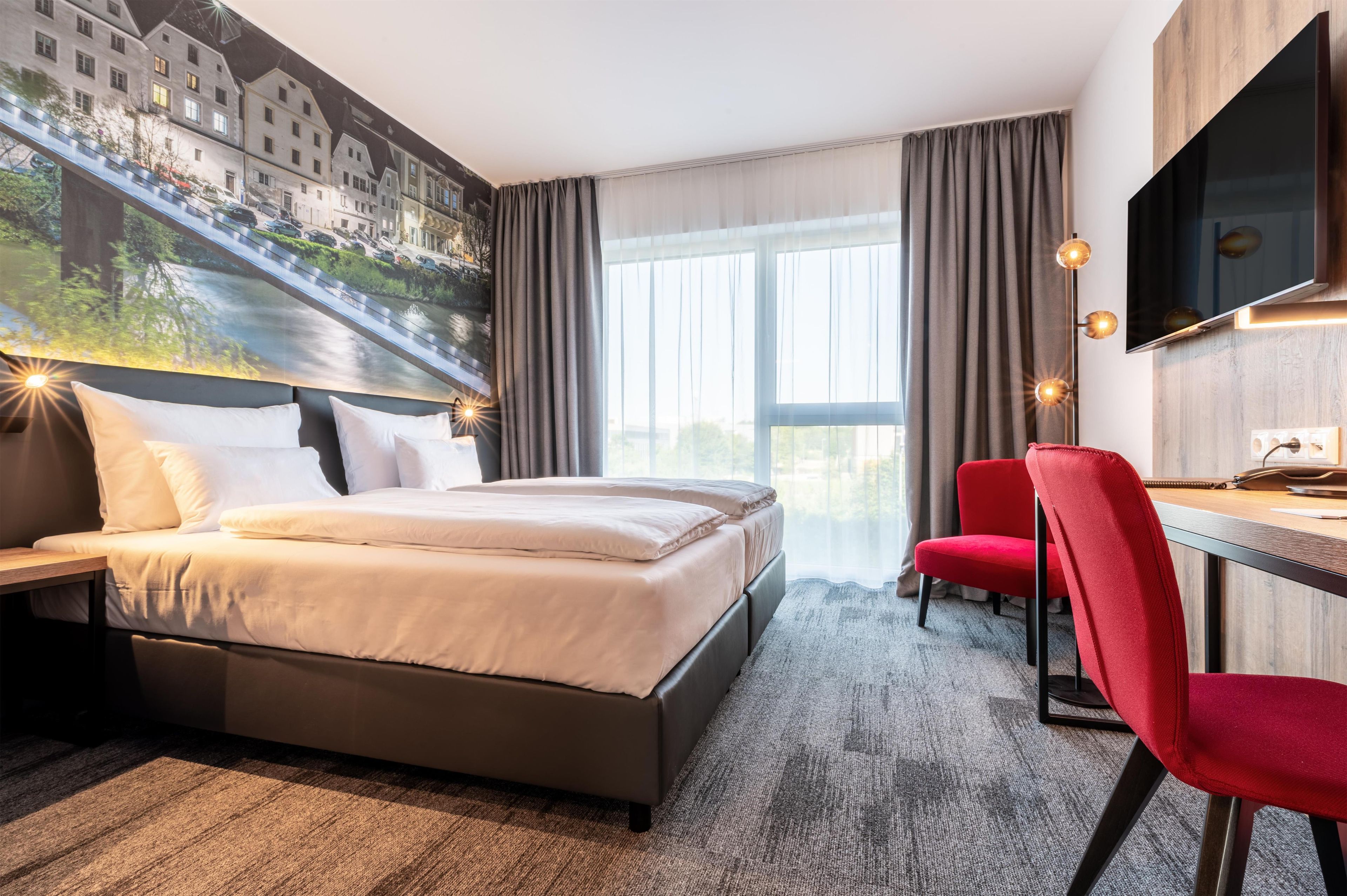 Habitaci ón de hotel con cama, escritorio, silla, televisor y un amplio ventanal con cortinas.