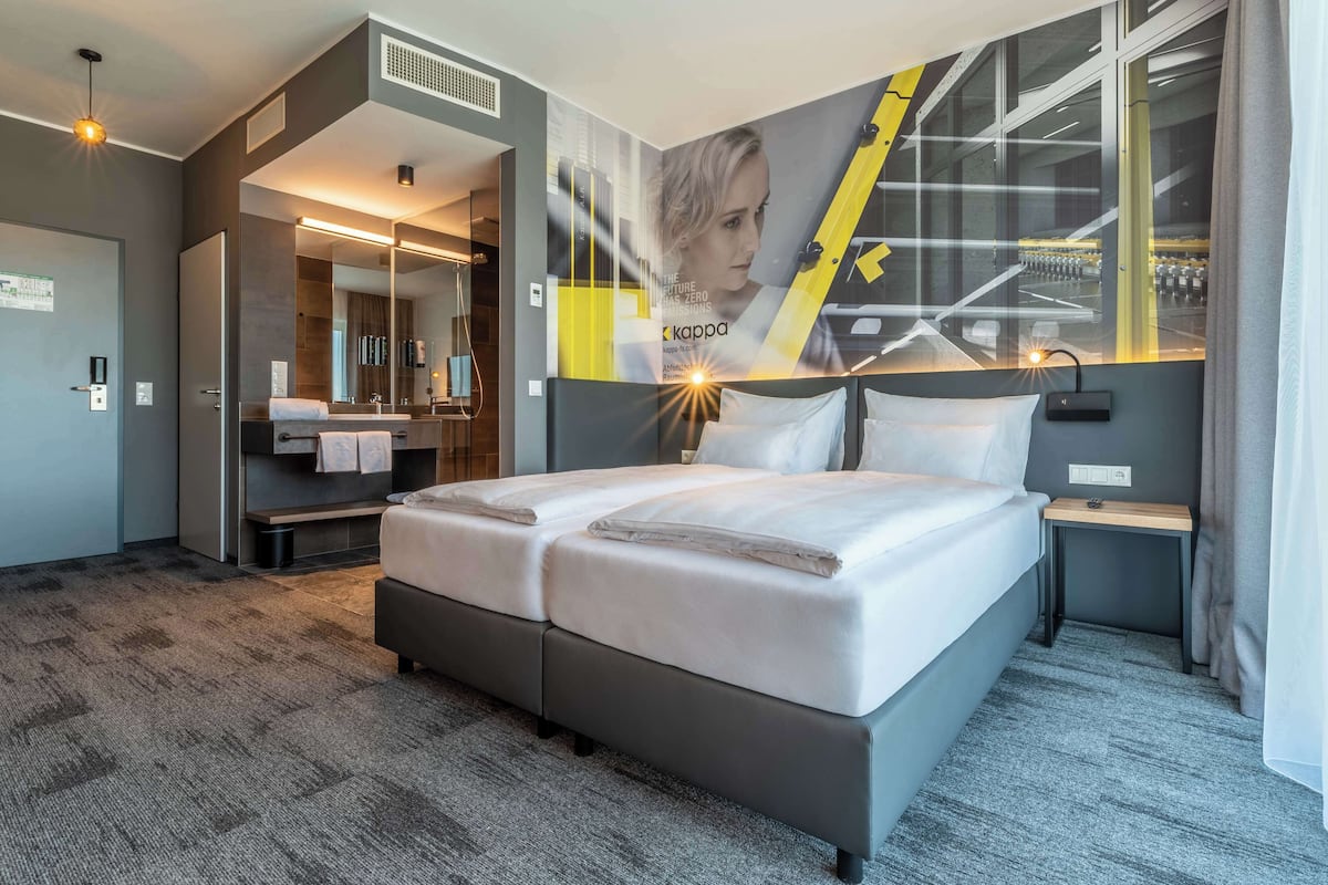 Ein modernes Hotelzimmer mit einem großen Bett, einem Badezimmer inklusive Spiegel und einer Wand mit einem Poster.