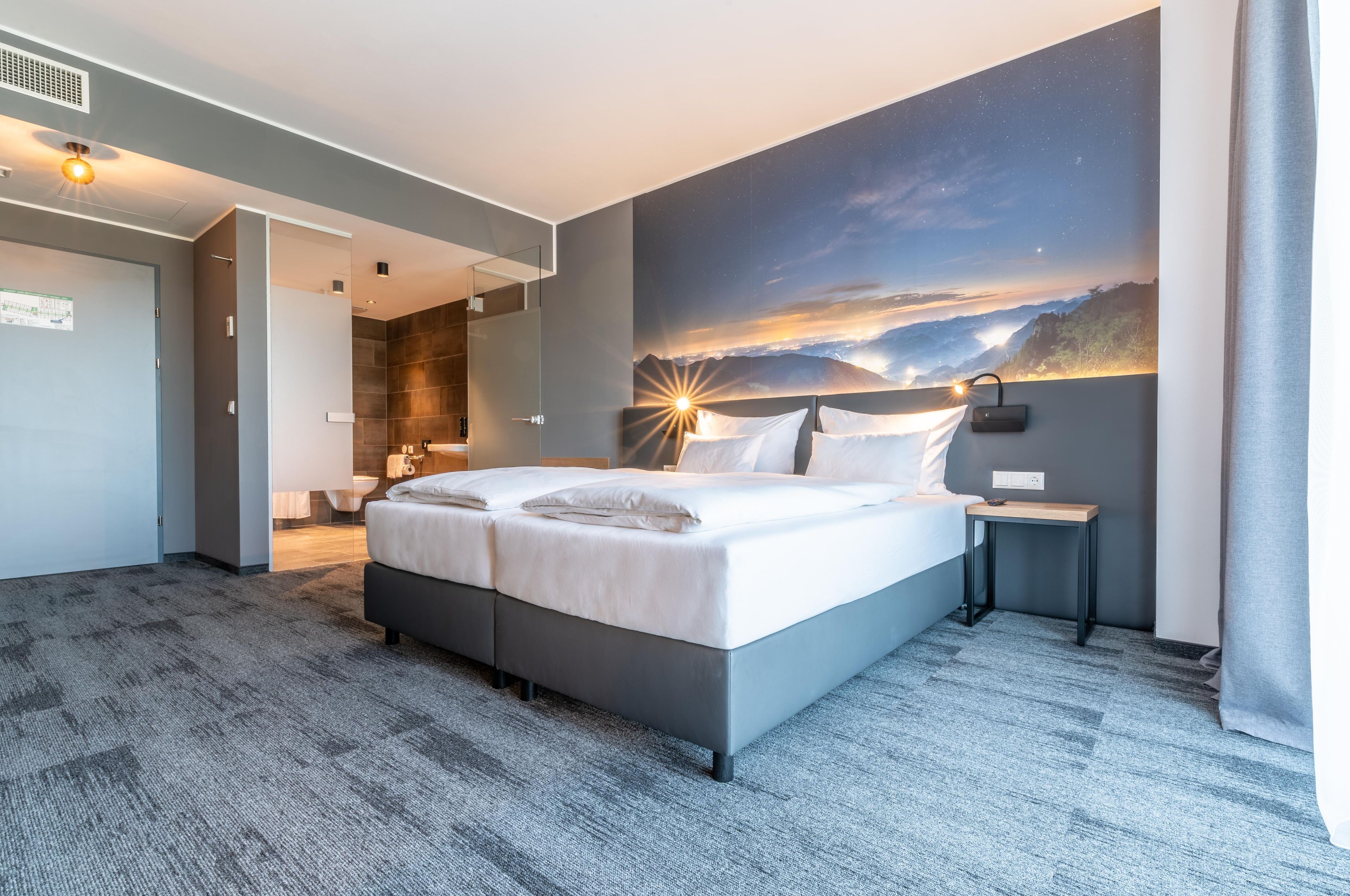 Una habitación de hotel moderna con una cama grande, un mural del cielo nocturno y vistas a un paisaje montañoso.