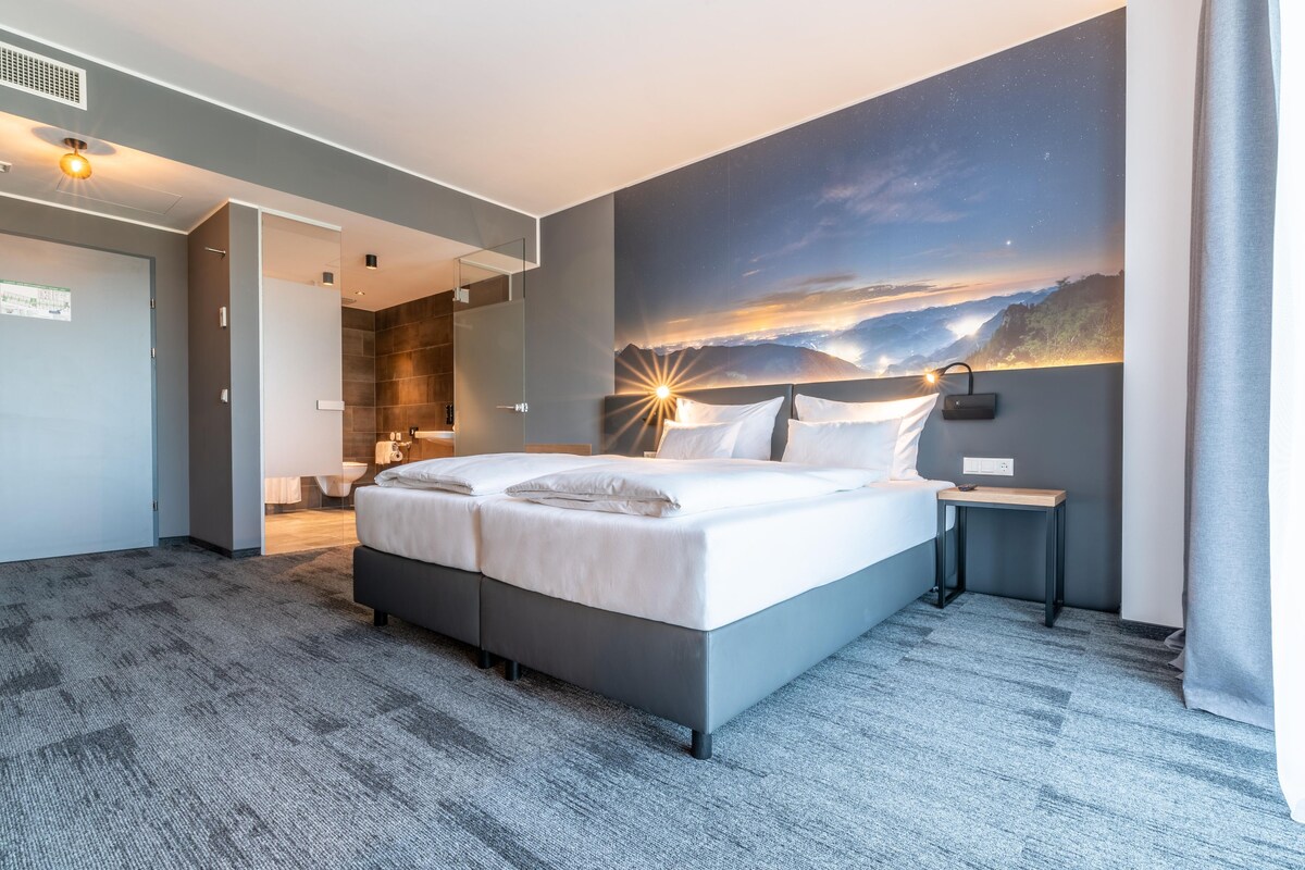 Une chambre d’hôtel moderne avec un grand lit, une fresque représentant un ciel étoilé et une vue sur un paysage montagneux.