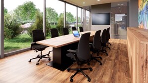 A conference room with a long table, black office chairs, and a large window with a view of greenery and a road.
