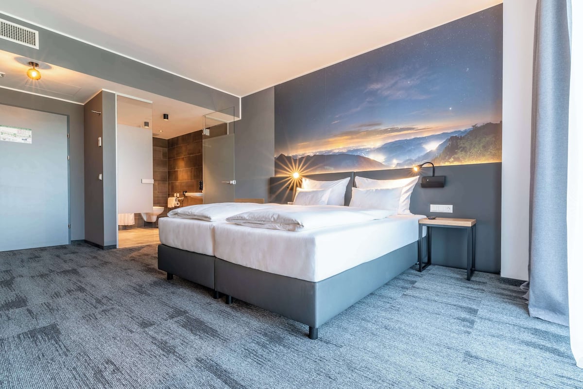 Ein modernes Hotelzimmer mit einem großen Bett, einem Wandgemälde mit Sternenhimmel und einem kleinen Nachttisch, mit Blick auf das Badezimmer.