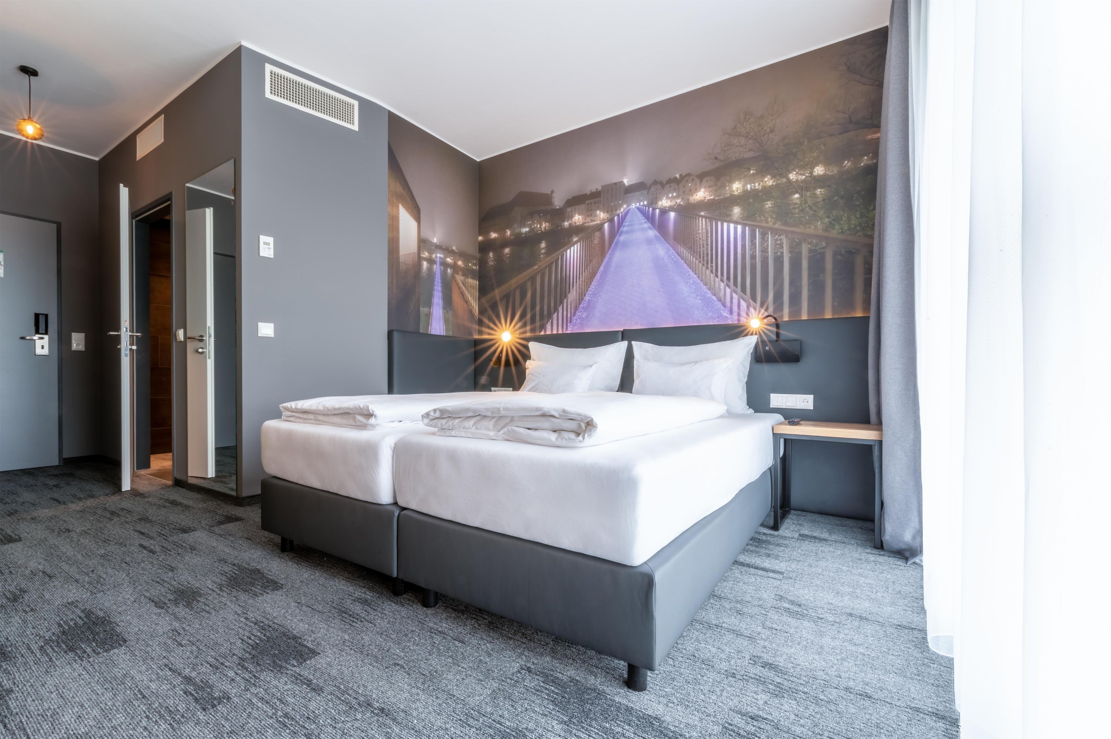 A hotel room with a large bed, a nightstand, a wall-mounted TV, and a door leading to another room.