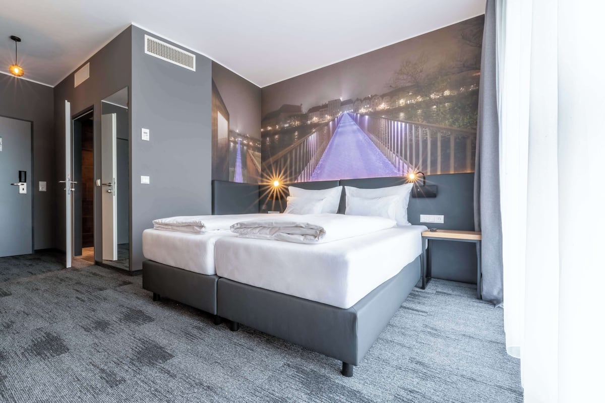 Ein Hotelzimmer mit einem großen Bett, einem Nachttisch, einem Spiegel und einem Wandgemälde einer Brücke.