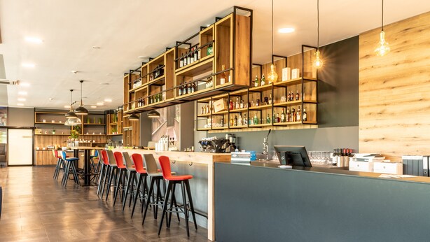 A modern bar area with wooden shelves, a long counter, and multiple bar stools. - Aiden by Best Western Stadtgut Hotel Steyr (Steyr)
