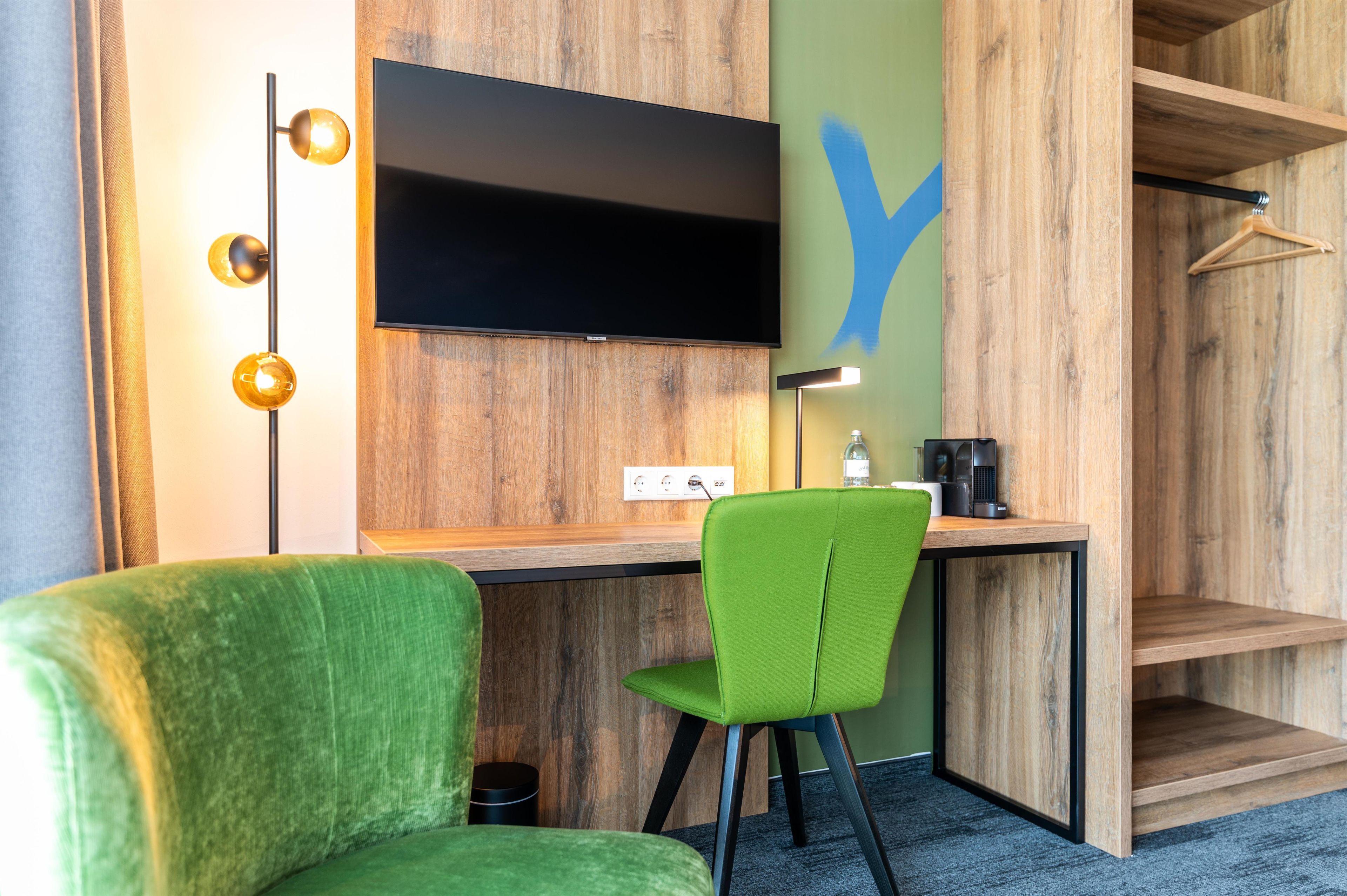 Habitación de hotel moderna con pared de madera, televisor de pantalla plana, una silla verde, un escritorio y un perchero.