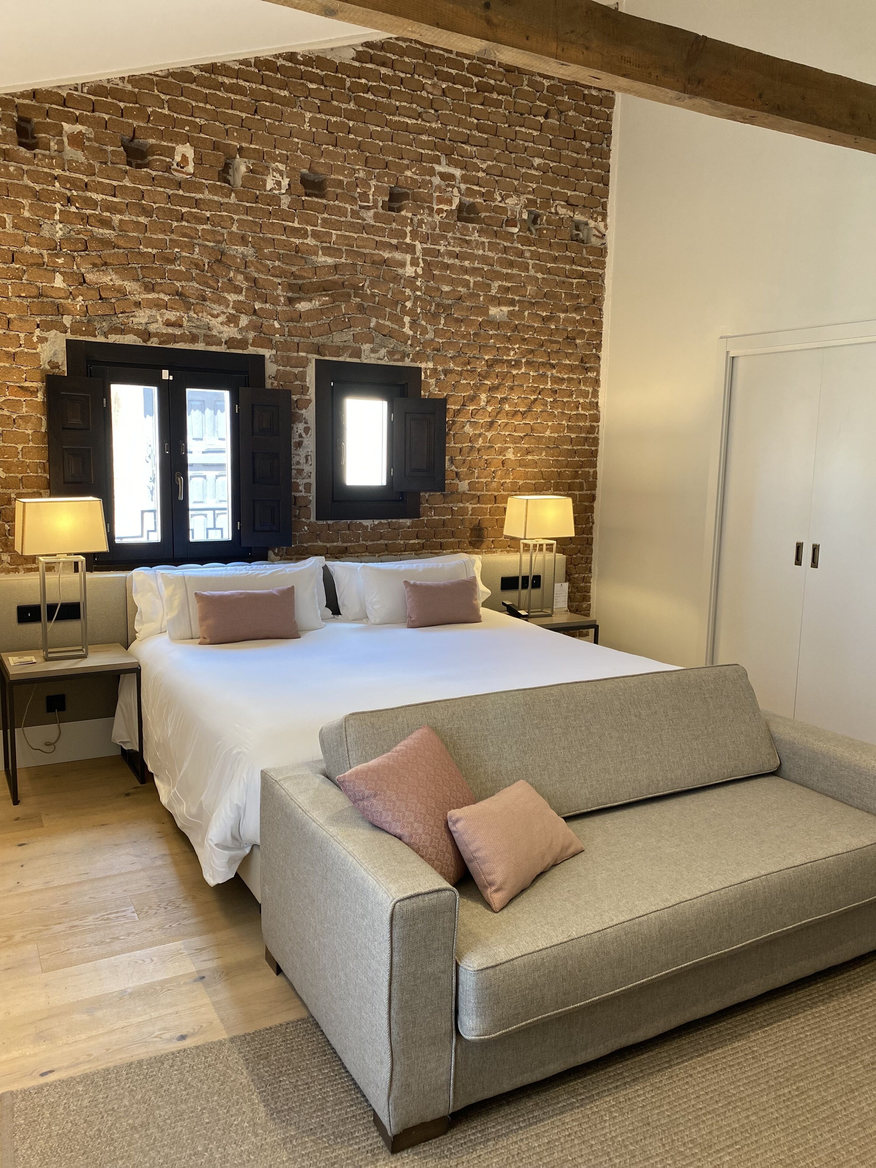 A bedroom with a brick wall, a bed with white bedding, two bedside tables with lamps, and a grey sofa.