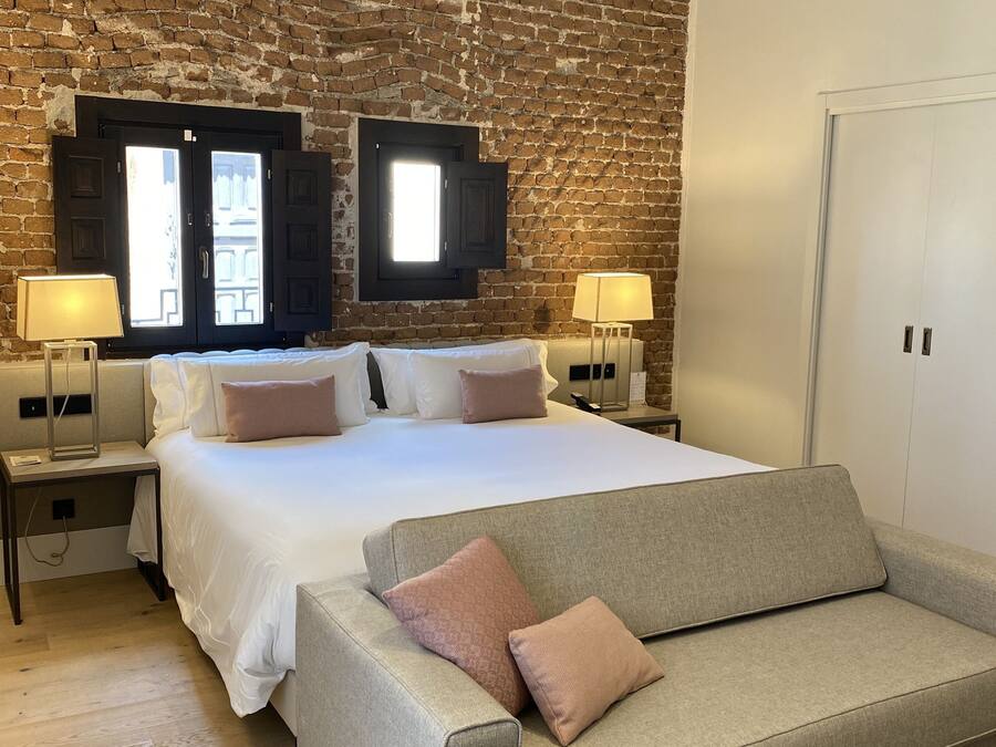 A bedroom with a brick wall, a bed with white bedding, two bedside tables with lamps, and a grey sofa.